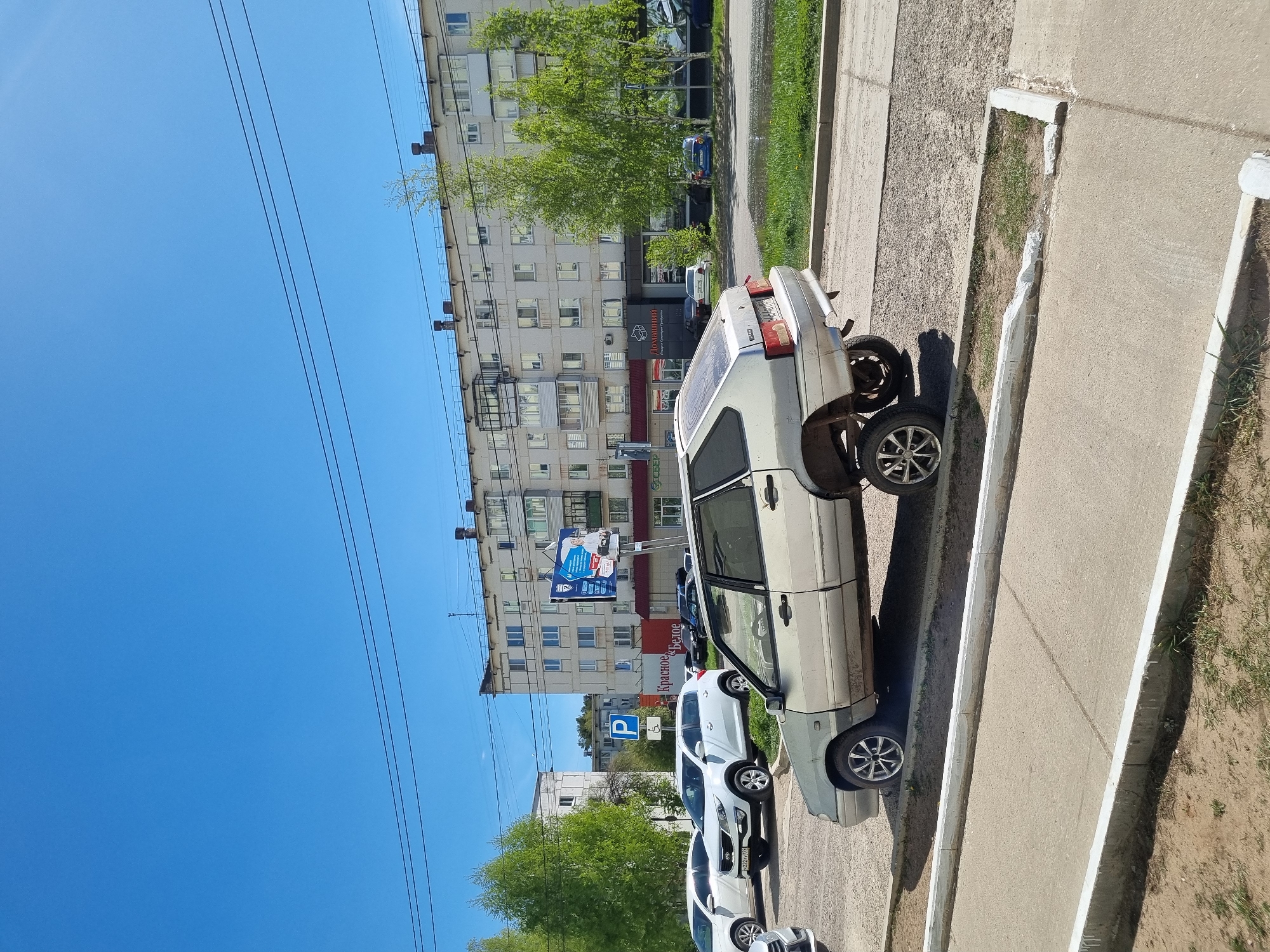 A daytime, street-level view in Chaikovsky, Russia, depicts a silver Lada parked along a concrete curb. Its right rear wheel is missing. A line of parked cars extends behind the Lada. Across the street, a multi-story apartment building is visible, featuring commercial signage in Russian. Power lines stretch across a clear blue sky.