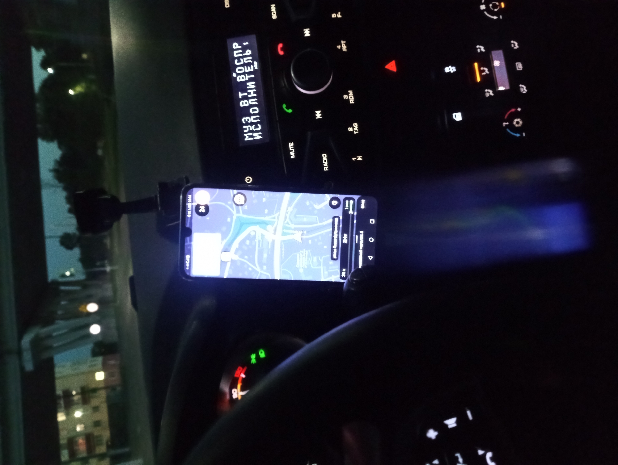 The image is a close-up, low-light, interior shot of a car at night. The focus is on the car's dashboard and a smartphone mounted on a car mount. The car's dashboard features various buttons and a digital display. The digital display shows Russian text, which translates to "MUSIC BLUETOOTH PERFORMER:". A map application is open on the smartphone, showing a route. The background is dark and blurred, suggesting the car is in motion, possibly on a highway or main road, with some distant lights from buildings and streetlights visible. The steering wheel is partially visible on the left side of the frame.