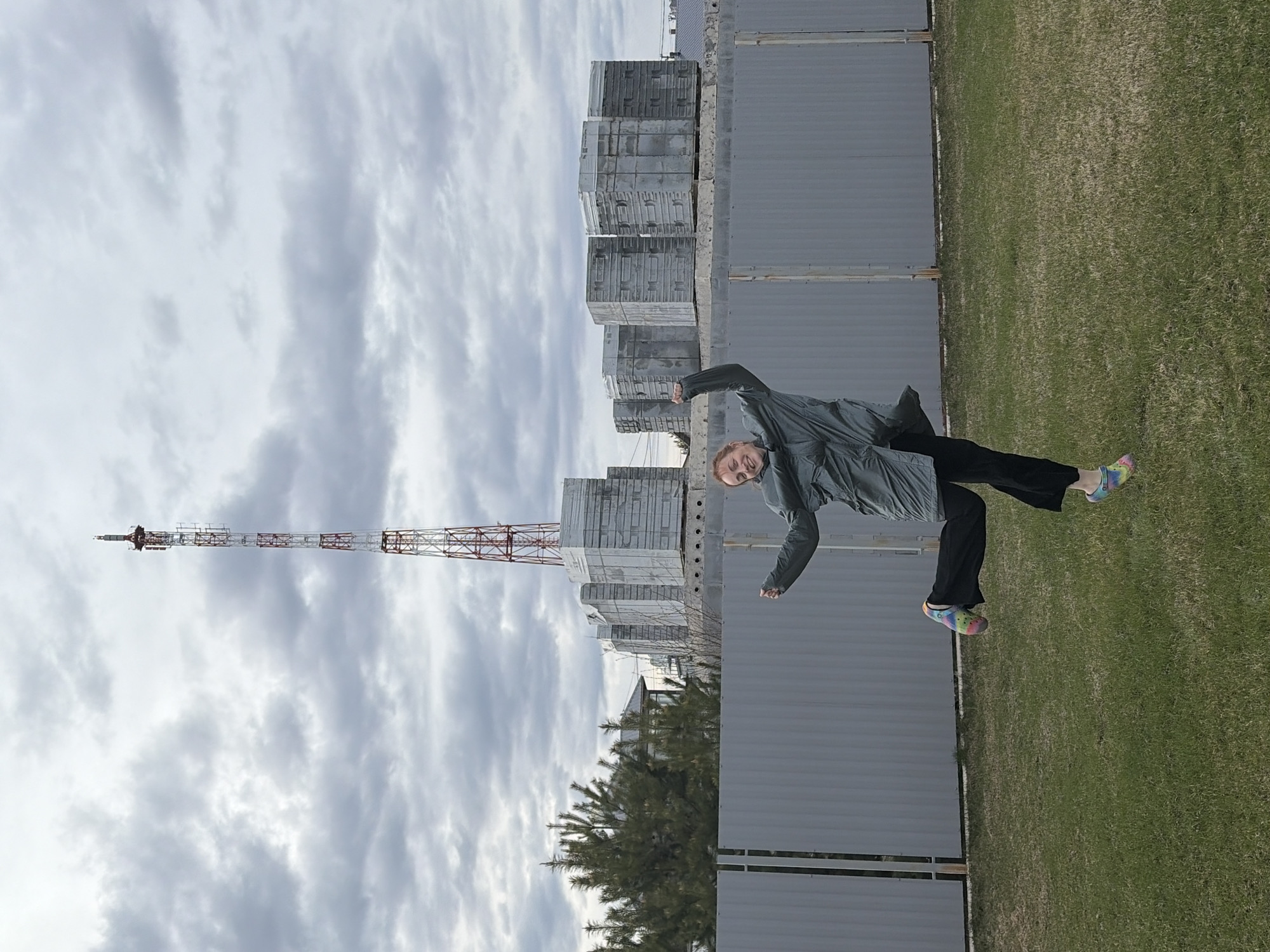 This image, set in Chaikovsky, Russia, captures a person striking a dynamic pose outdoors under a cloudy sky. The main subject is a young individual, positioned slightly right of center, wearing a dark grey jacket or raincoat, black trousers, and distinctive, brightly colored tie-dye clogs. They are smiling broadly at the camera, conveying a cheerful and playful mood, as they lean forward with arms slightly bent, almost as if in mid-jump or dance.
The scene features a grassy foreground, transitioning into a tall, grey corrugated metal fence that spans horizontally across the middle of the frame. Behind this fence, several large stacks of what appear to be concrete blocks or industrial materials are visible, suggesting an industrial or construction area. In the background, against a vast, overcast sky filled with light and dark grey clouds, stands a prominent, tall lattice telecommunications tower, painted in alternating red and white sections. Some green foliage, possibly a pine tree, peeks out from behind the fence on the left side. The weather appears to be a cloudy daytime, and the overall environment leans towards a utilitarian or semi-industrial landscape. There is no visible text in the image.