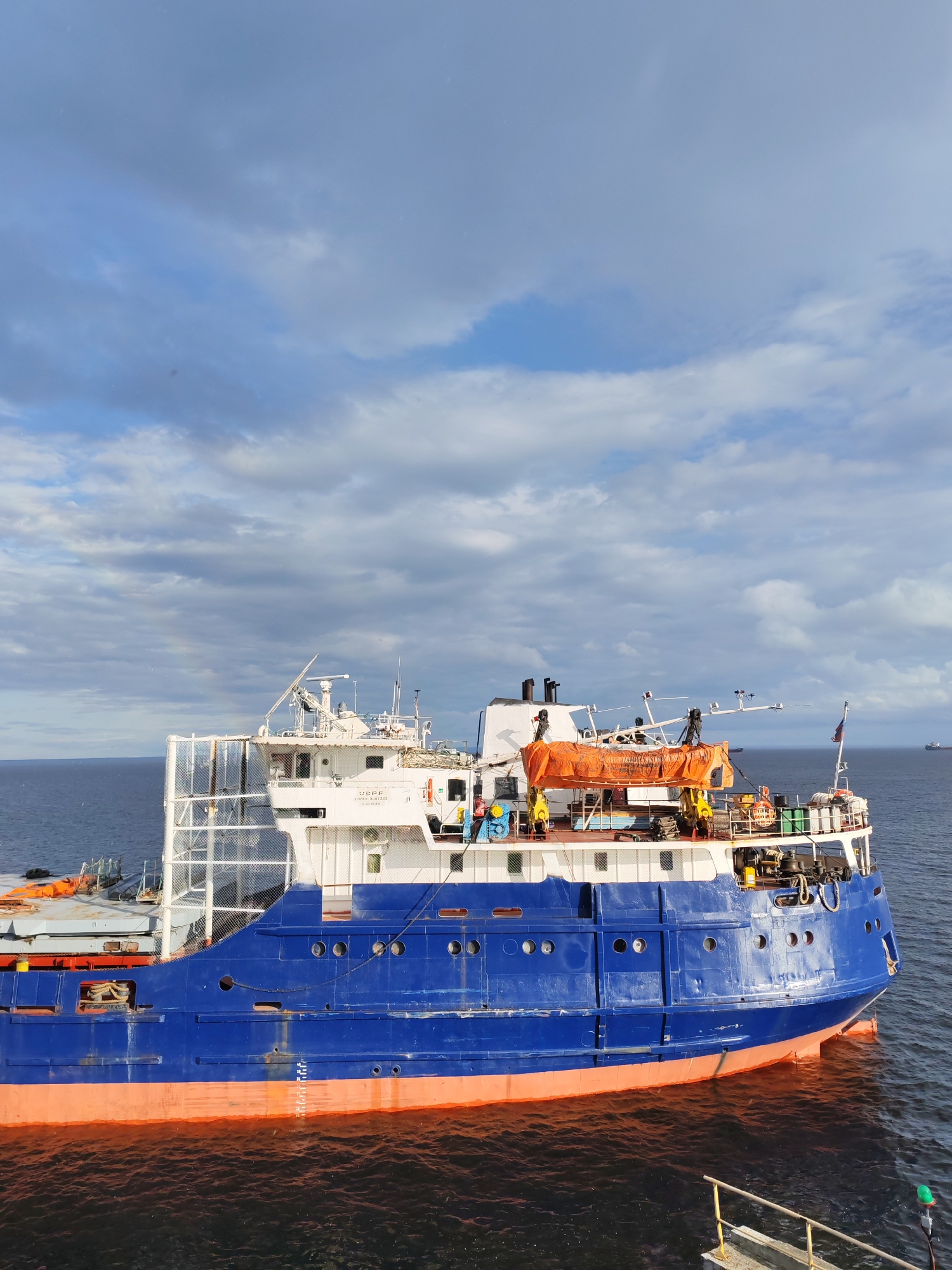 An overhead, close-up view shows the starboard side of a blue and orange cargo ship docked in the city of Shcheleiki, Russia. The ship is partially submerged in dark blue water, with a faint rainbow visible in the distance. The ship's superstructure is white, with multiple decks and windows. An orange life raft is visible on the top deck. The sky is cloudy and overcast. The overall scene suggests a day with inclement weather.