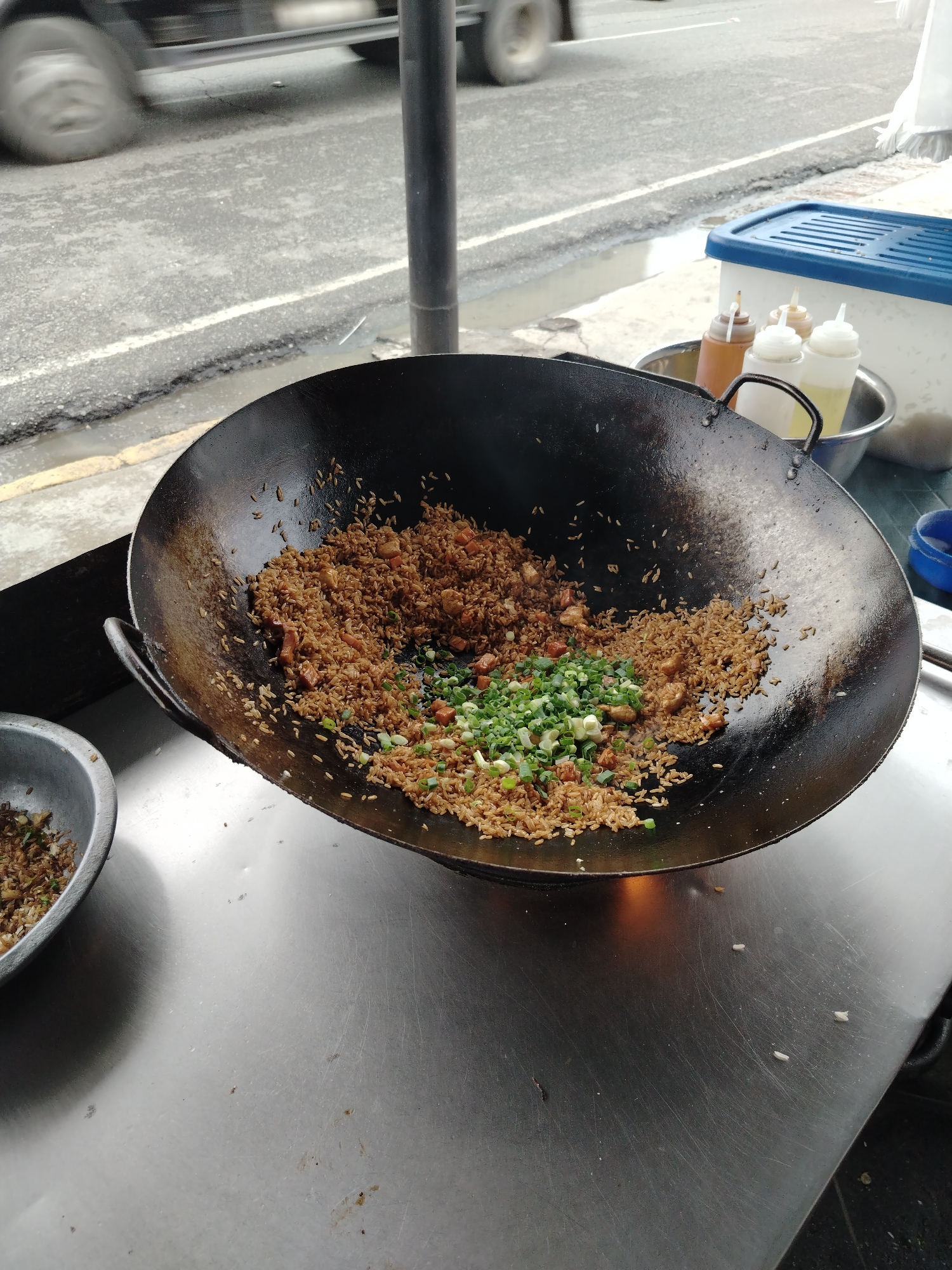 This image captures an outdoor street food scene, likely a vendor preparing fried rice, with a bustling road in the background. The central object is a large, well-used black wok, filled with brownish-orange fried rice. The rice appears to be seasoned with soy sauce or similar ingredients and contains small cubes, possibly chicken, pork, or vegetables like carrots. A vibrant pile of freshly chopped green onions rests on top of the rice, indicating a final addition to the dish.
To the left, a smaller metal bowl also contains some fried rice, possibly a serving bowl or for holding ingredients. The food preparation takes place on a metallic counter, which shows signs of wear and tear, including scratches and minor residue. In the upper right background, several squeeze bottles, likely holding sauces or oils for cooking or serving, are gathered in a metal bowl beside a white container with a blue lid.
Beyond the food stall, a paved road stretches across the background. A vehicle, likely a truck or bus, is captured in motion on the road, evident by the blur of its wheels and body, suggesting a dynamic and busy environment. The road surface appears somewhat damp in places. A dark, vertical pole stands between the road and the food stall. The setting conveys a typical daytime street food operation in an urban or semi-urban area, consistent with locations like Tinaquillo, Venezuela. There is no visible text in the image.