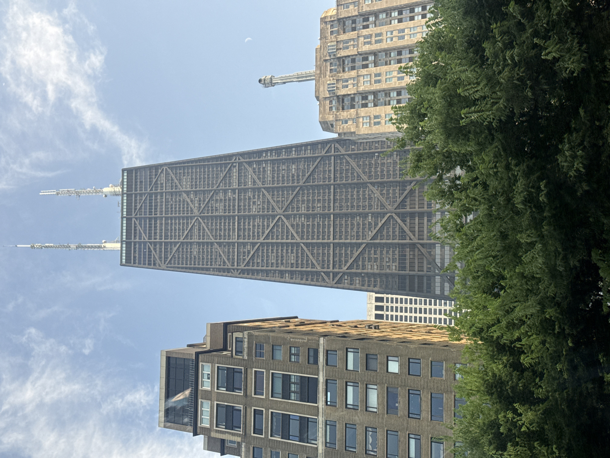 The image shows a low-angle view of several buildings in Chicago, Illinois, United States of America. The most prominent structure is the John Hancock Center, a skyscraper with a distinctive X-braced exterior and two antennas on its roof. To the left of the John Hancock Center is a brown brick building with many windows, and to the right, partially obscured by trees, is another stone-clad building with a unique spire-like structure on its roof, resembling the historic Carbide & Carbon Building. In the background, a crescent moon is visible in the hazy sky. Lush green trees fill the foreground, obscuring the lower portions of the buildings. The overall scene depicts an urban landscape on a clear day with a slight atmospheric haze.