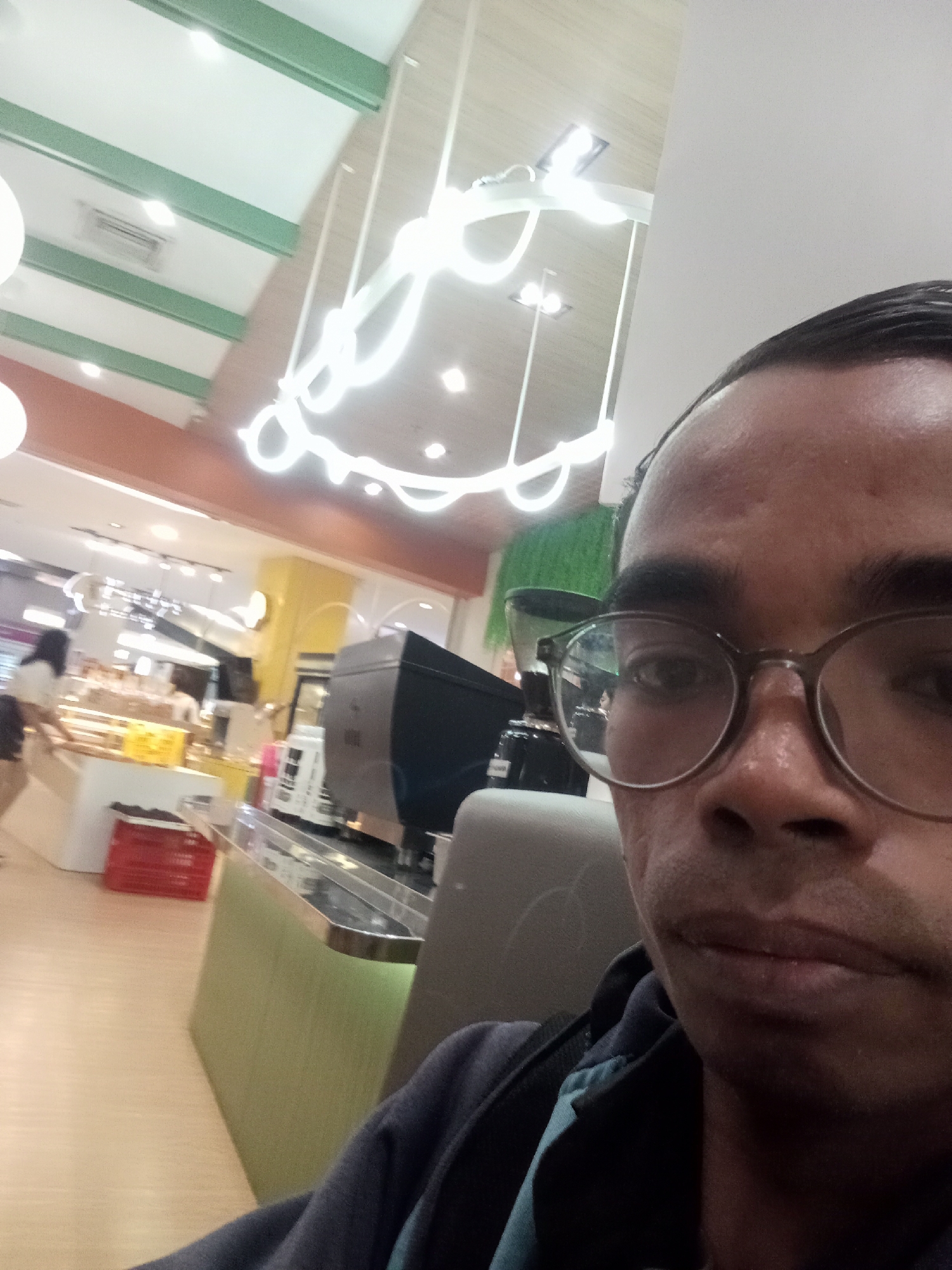 The image is a selfie taken by a person wearing glasses in what appears to be a brightly lit cafe or restaurant. In the background, there are tables, chairs, and a counter with food items. The person's face is the primary focus of the image. There are no discernible landmarks or clues that specifically identify the location as Surabaya, Indonesia. The scene suggests a casual indoor environment, possibly a public eatery.