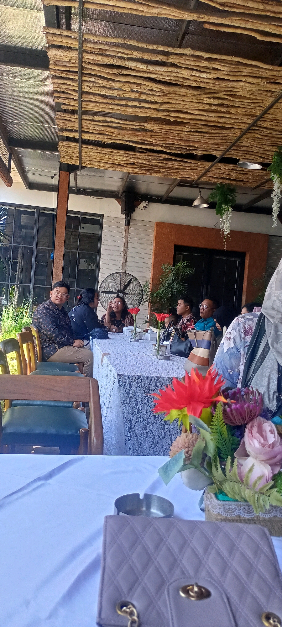 The image depicts an outdoor gathering of approximately seven individuals seated at a long table covered with a white lace tablecloth. The setting appears to be a restaurant or cafe with a rustic, natural aesthetic, featuring a ceiling constructed of interwoven branches and exposed metal beams.
The individuals are dressed in a mix of formal and casual attire. On the left side of the table, a man in a patterned dark shirt and light-colored pants is looking towards the camera. Next to him, a woman in a dark top is engaged in conversation. Further down the table, several other individuals are visible, some in conversation and others looking around. A baby is also present, seated on the lap of an adult.
On the table, there are several flower arrangements in vases, as well as what appears to be a packet of cigarettes and an ashtray. In the foreground, a light purple quilted purse with gold hardware is placed on a white tablecloth. The background shows a wall with a large fan and some greenery.
The overall atmosphere suggests a social event, possibly a family gathering or a business meeting, taking place in a semi-open, shaded area. The presence of plants and natural materials in the construction of the venue indicates a connection to nature. There is no specific indication within the image to pinpoint the exact location within Desa Ponokawan, Indonesia, beyond the general context of it being a public establishment.