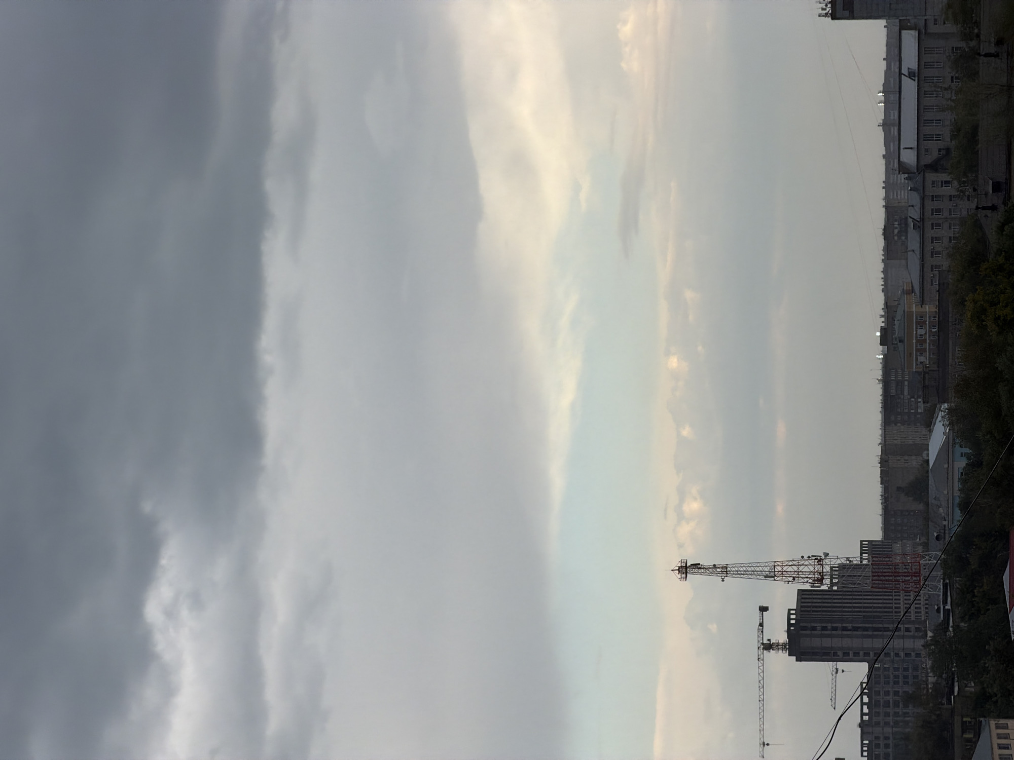 The image depicts an overcast sky above a city skyline, likely Pavlodar, Kazakhstan. In the foreground, a tall, red and white lattice communications tower stands prominently. To its left, several construction cranes are visible near unfinished buildings, suggesting ongoing development. The middle ground features a collection of residential and industrial buildings. The background is dominated by a dense layer of dark, heavy clouds, hinting at impending weather. The overall impression is one of an industrial or developing urban landscape under a subdued, cloudy sky.