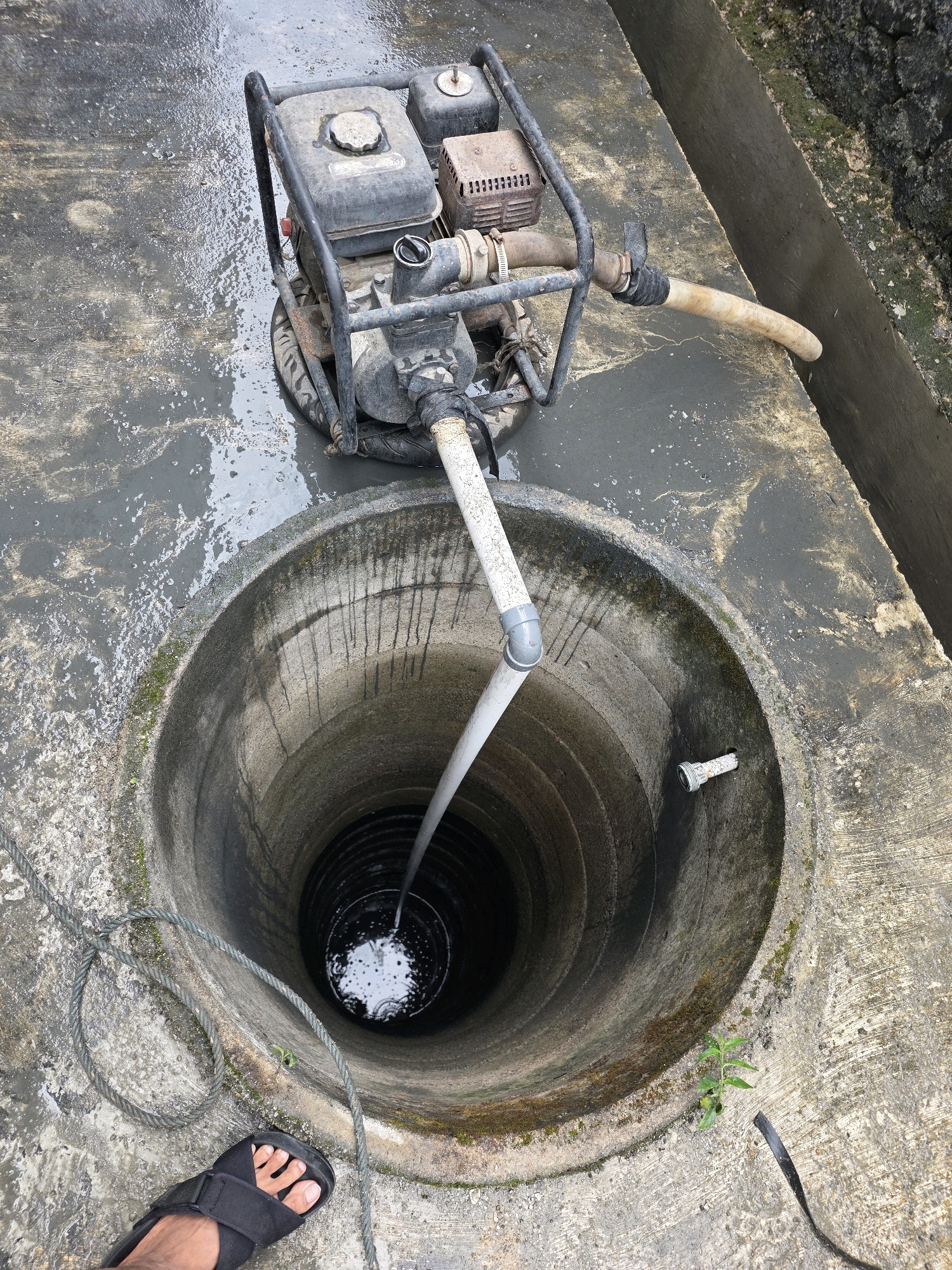 In the city of Tanjung Pinang, Indonesia, a water pump is positioned on a wet concrete surface next to a deep, circular well. The pump, a portable gasoline-powered unit, is connected to a white pipe that extends down into the well. A hose from the pump directs water away from the well. The concrete area around the well appears weathered and there is a small plant growing nearby. A rope lies coiled on the ground. A person's foot wearing a black sandal is visible in the lower left corner of the image. The scene suggests a water extraction or drainage operation.