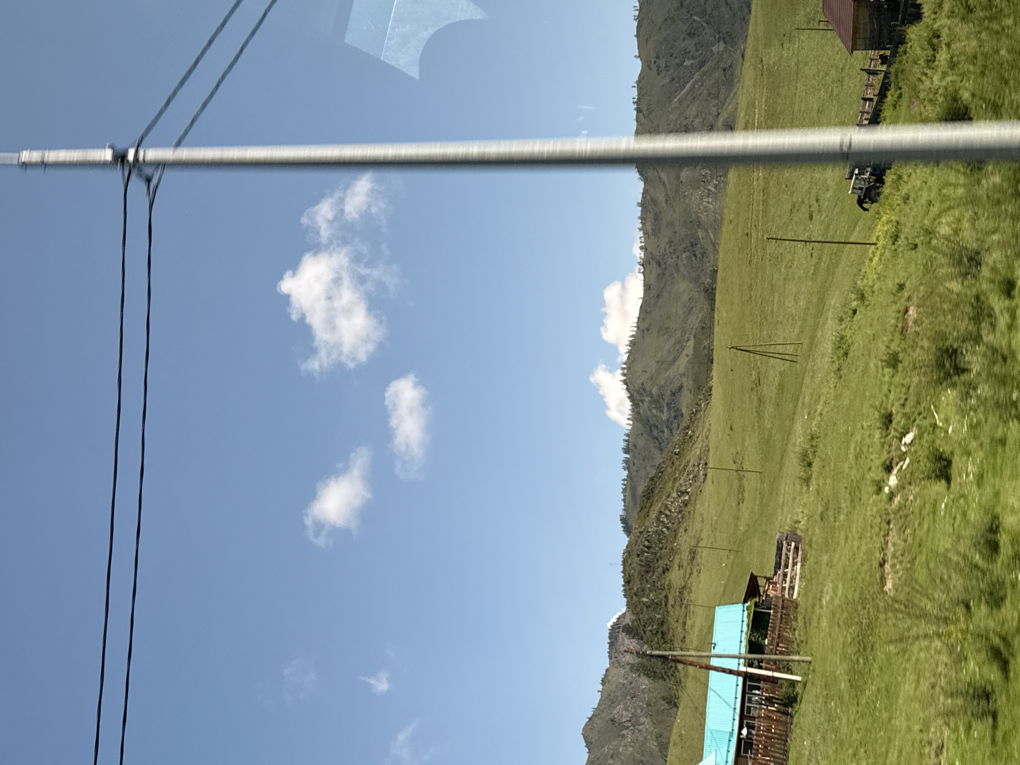 This is a photograph taken from a moving vehicle, likely a car, looking out at a rural landscape in the vicinity of Tuekta, Russia. The scene is dominated by a vast, green, rolling pasture under a bright blue sky with scattered white clouds. In the mid-ground, a small wooden house with a distinctive turquoise roof and a wooden fence is visible on the left side of the frame. A wooden pole, likely a utility pole, stands in front of the house. Further into the field, to the right of the house, a blue tractor is parked on a slight incline. In the background, rugged, tree-lined mountains recede into the distance. Power lines are strung across the top of the image, and a tall, metallic utility pole runs vertically along the right side of the frame, obscuring part of the view. The overall impression is one of a serene, natural environment in a remote, possibly agricultural, setting.