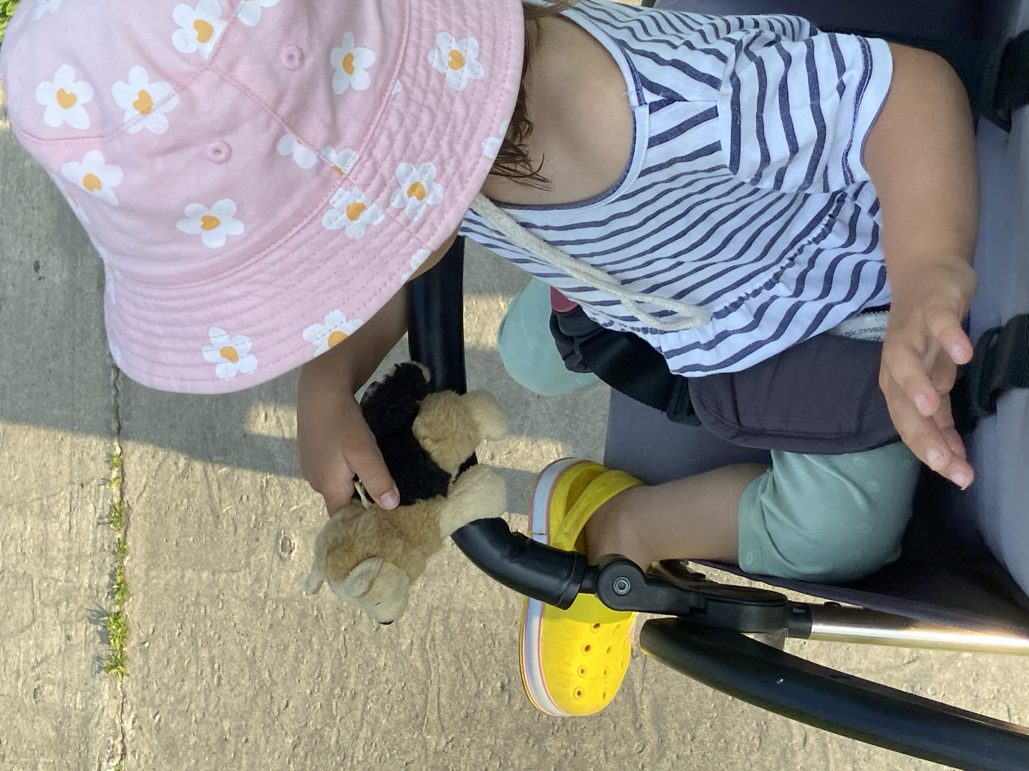 A young child, wearing a pink bucket hat with white daisy patterns and a blue and white striped shirt, is seated in a stroller. The child's left leg, clad in a pale green outfit, is visible, along with a yellow Croc shoe. The child holds a small stuffed dog toy. The stroller has black handles and a dark gray seat. The ground appears to be a paved surface with some cracks and small plants growing through. The scene is outdoors, in daylight, with shadows cast on the ground, suggesting a sunny day. The location is identified as the city of Tyumen, Russia.
