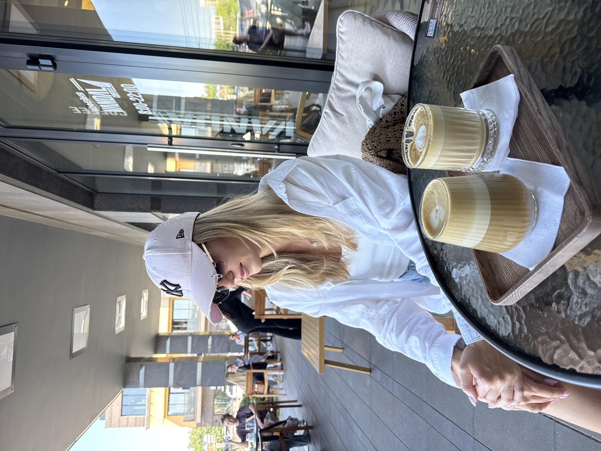 The image shows a young woman sitting at a glass table outdoors, possibly at a cafe. She is wearing a white button-down shirt, a white baseball cap, and sunglasses. Her blonde hair is visible beneath the cap. Two glasses of coffee with latte art are on a wooden tray on the table in front of her, along with a white napkin. Her hands are clasped on the table. In the background, other patrons are seated at tables, and a building with large windows is visible. The reflection in the window shows text, possibly the name of the establishment and a phone number. The scene appears to be during the daytime, with bright lighting.