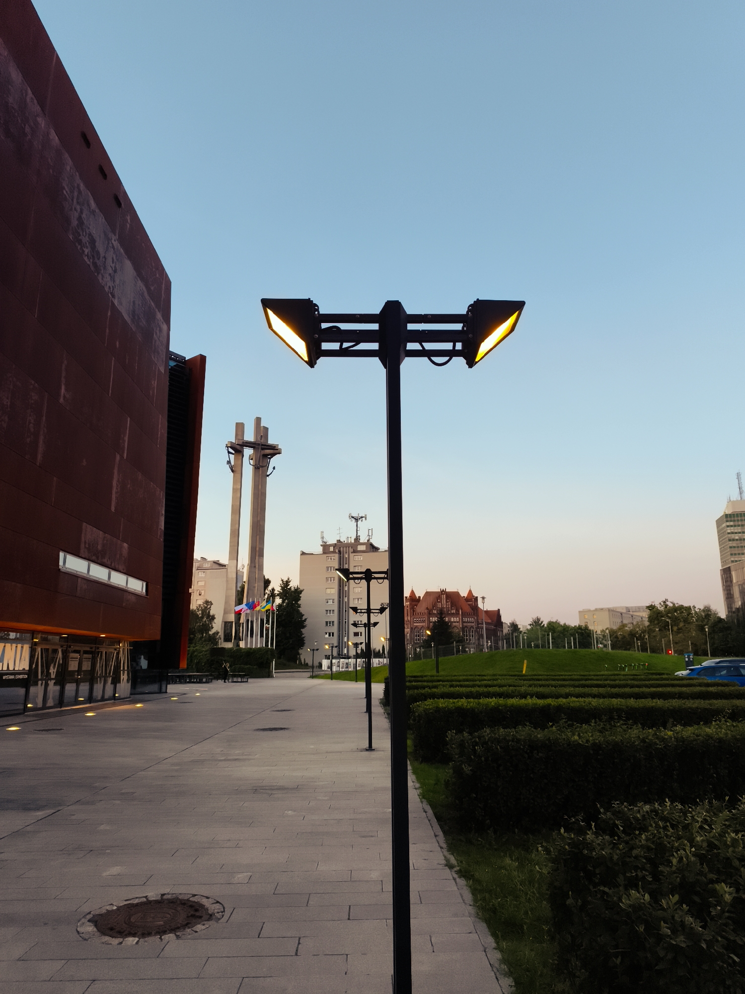 The image depicts an outdoor scene at dusk in Gdańsk, Poland, specifically at the European Solidarity Centre. In the foreground, a modern building with a reddish-brown facade and large glass doors is visible on the left. Adjacent to it is a paved walkway with square paving stones. A lamppost with two bright, angled lights stands prominently in the center of the walkway, casting a warm glow. In the background, several other lampposts line the path. Further back, a large monument with three cross-shaped structures is visible, along with several flags. Beyond that, various buildings, including residential blocks and a distinct red-bricked building, dot the landscape. The sky is a clear gradient of blue to pale pink, indicating twilight. A meticulously maintained hedge borders the walkway on the right, with a patch of green grass beyond it. A blue car is parked on the right side of the image. The overall atmosphere is serene and contemporary, capturing the urban environment of Gdańsk at the end of the day.