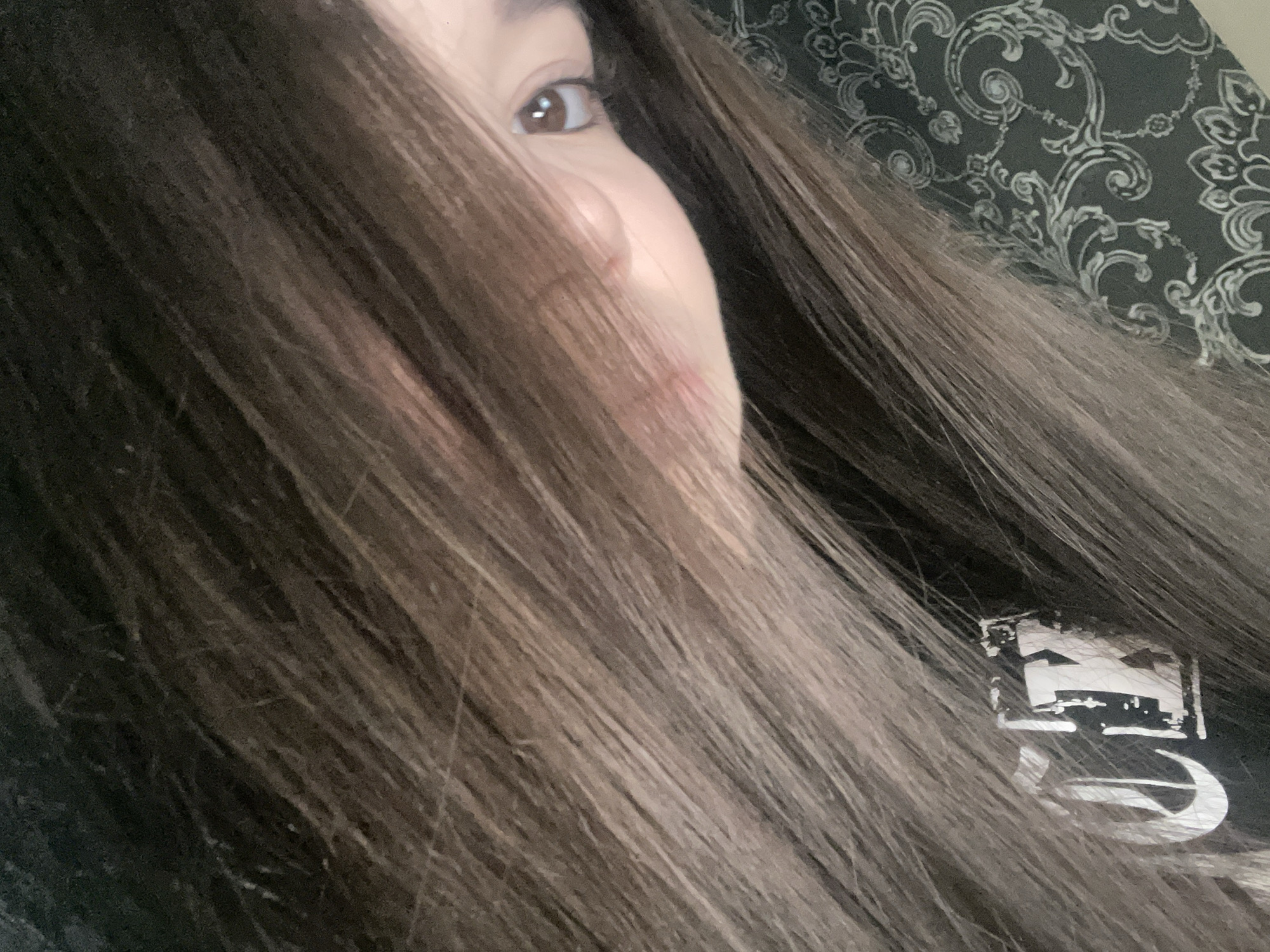 The image displays a close-up, angled portrait of an individual, likely female, captured indoors. The subject's face is predominantly obscured by long, dark brown hair, which appears to have lighter brown or auburn streaks and sweeps across the majority of the frame from the upper left to the lower right. A single dark brown eye with a distinct catchlight is visible on the upper right quadrant, looking directly towards the viewer. A portion of the nose and the left side of the mouth/cheek area, exhibiting light skin pigmentation, are also visible beneath the hair. The subject is wearing a dark, possibly black, garment with a partially visible white logo or lettering on the left side, which includes characters resembling an ornate 'C' followed by an 'X'. In the lower right corner, a dark patterned fabric, possibly a pillowcase or upholstery, with a white or light grey intricate floral or paisley design is present. The specific location within Chaikovsky, Russia, cannot be determined from the visual evidence provided.