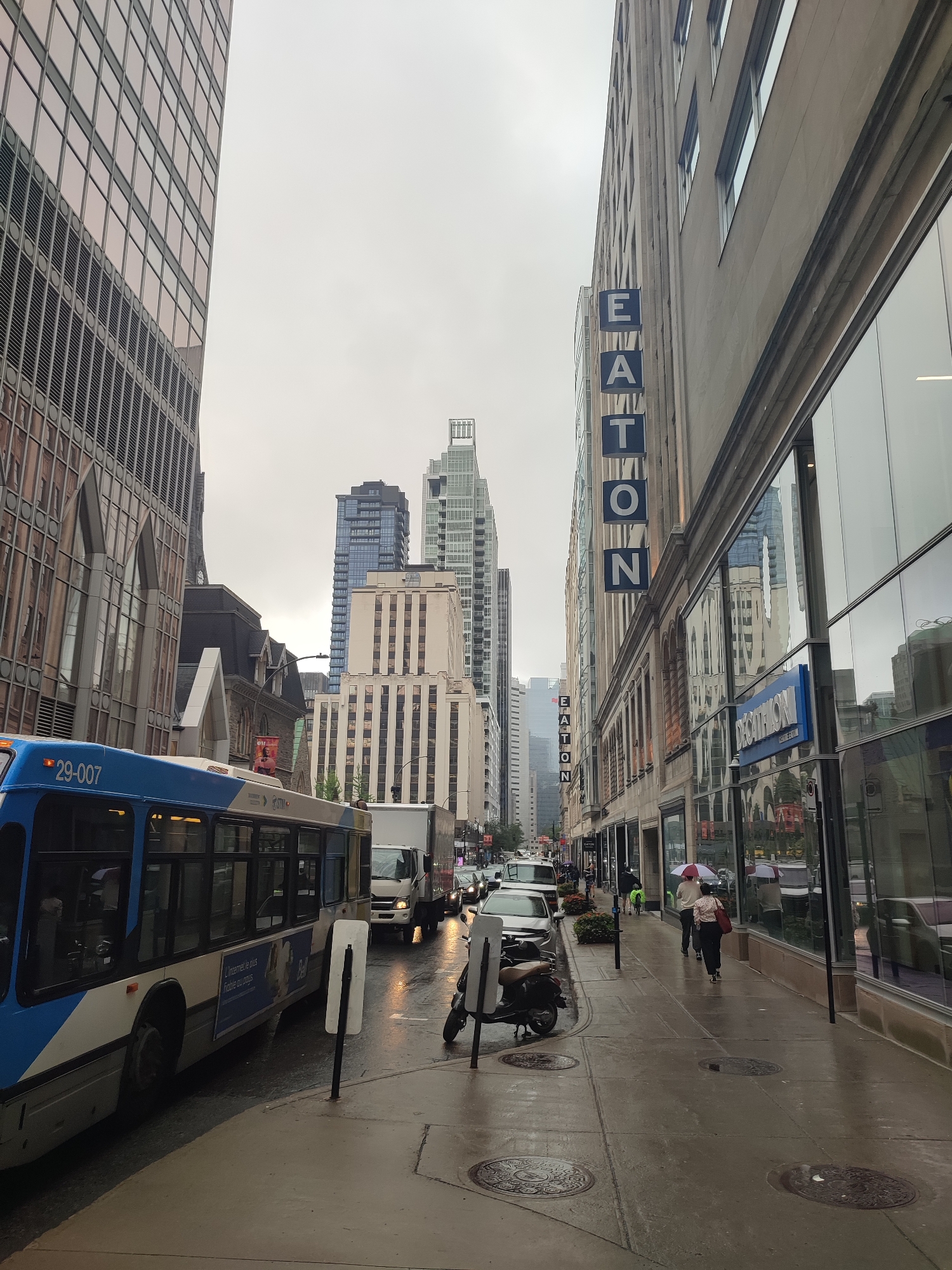 This image depicts a street scene in Montreal, Canada, with a focus on a bus, surrounding buildings, and pedestrians on a wet sidewalk. The perspective is from street level, looking down a city street lined with tall buildings.
In the foreground, a blue and white public transit bus with the number "29-007" is prominently featured on the left side of the frame. The bus appears to be in motion or stopped in traffic. To the right of the bus, a wet sidewalk is visible, with several pedestrians walking. One person is walking with a red and white umbrella. Other pedestrians are visible further down the sidewalk. A scooter is parked on the sidewalk near the bus.
The buildings lining the street are a mix of architectural styles. On the left, a modern building with a glass facade reflects the sky and other structures. Further down the street on the right, a building features a prominent blue sign with white lettering that reads "EATON," indicating a commercial establishment. Below the "EATON" sign, another blue sign displays "DECATHLON" in white lettering. The architecture suggests a commercial district.
The street is wet, indicating recent rain, and reflects the surrounding lights and buildings. Traffic is present, with cars and a truck visible in the middle ground, suggesting a busy urban environment. The sky is overcast, contributing to the subdued lighting of the scene. In the background, more skyscrapers and urban structures are visible, reinforcing the city setting. The overall atmosphere is that of a typical urban day in Montreal, possibly during a damp or rainy period.