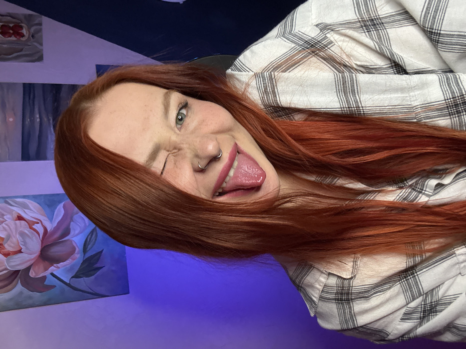 A close-up, informal portrait depicts a young female subject indoors. The subject has long, reddish-auburn hair, a fair complexion with visible freckles, and a silver-colored septum ring. She is wearing a white and black plaid shirt. Her right eye is closed in a wink, while her left eye, with a green iris, is open and directed towards the camera. Her mouth is open, revealing an extended tongue and deep pink lipstick. The background consists of a light purple wall illuminated by a blue light source from the left. Visible on the wall are three paintings: a large vertical painting of a pink peony on a blue background (left), a horizontal seascape with a setting sun/moon (center-right), and a smaller vertical painting depicting red fruit (far right). A dark blue angular panel or section is also visible on the far right. The scene is stated to be located in Rostov-on-Don, Russia.
