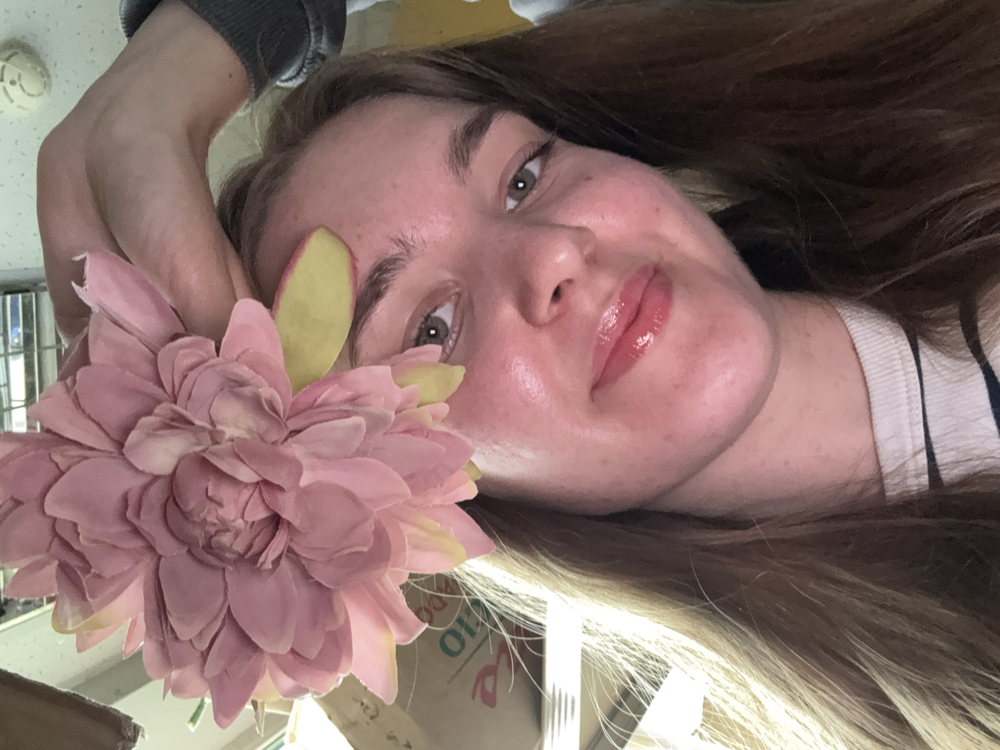 A person with long brown hair is lying on their back, facing the camera. They are holding a large pink flower near their face. The person is fair-skinned with light eyes and glossy lips. The background is cluttered, including a ceiling fixture, boxes, and a window. There is no verifiable evidence in the image that definitively places the location in the city of Perm, Russia.