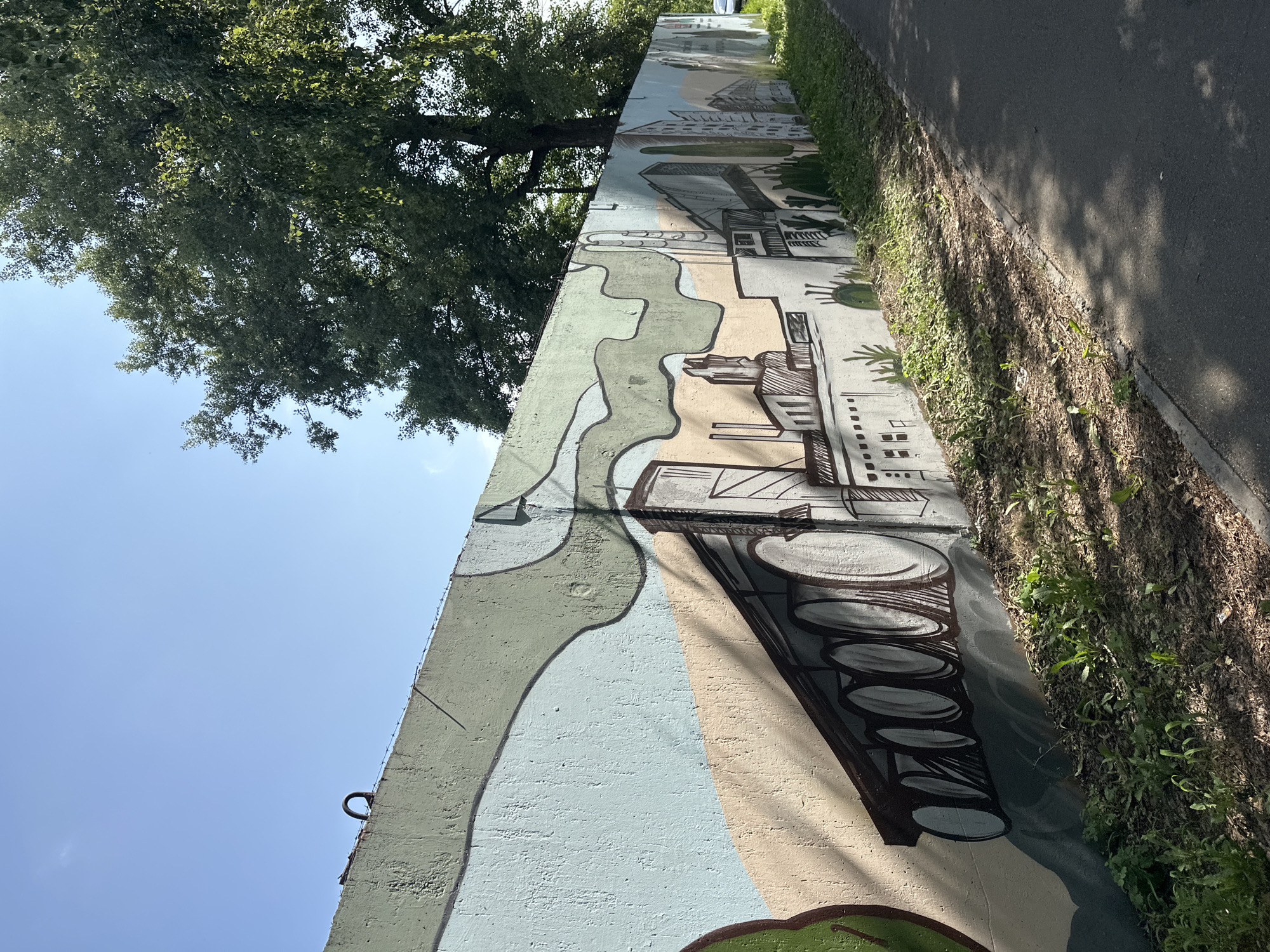 The image depicts a concrete wall adorned with a mural depicting an industrial landscape. The mural features stylized buildings, smoke stacks, and what appears to be a large train or vehicle with multiple wheels. The artwork is rendered in muted colors with bold black outlines. To the right of the wall is an asphalt path or road, bordered by a strip of grass and vegetation. A large tree with dense foliage casts shadows across the wall and the path. The sky is clear and blue. The wall itself shows signs of age, with some rough texture visible. A metal loop and barbed wire are present along the top edge of the wall. This mural is located on a retaining wall along the Ob River embankment in Biysk, Russia.