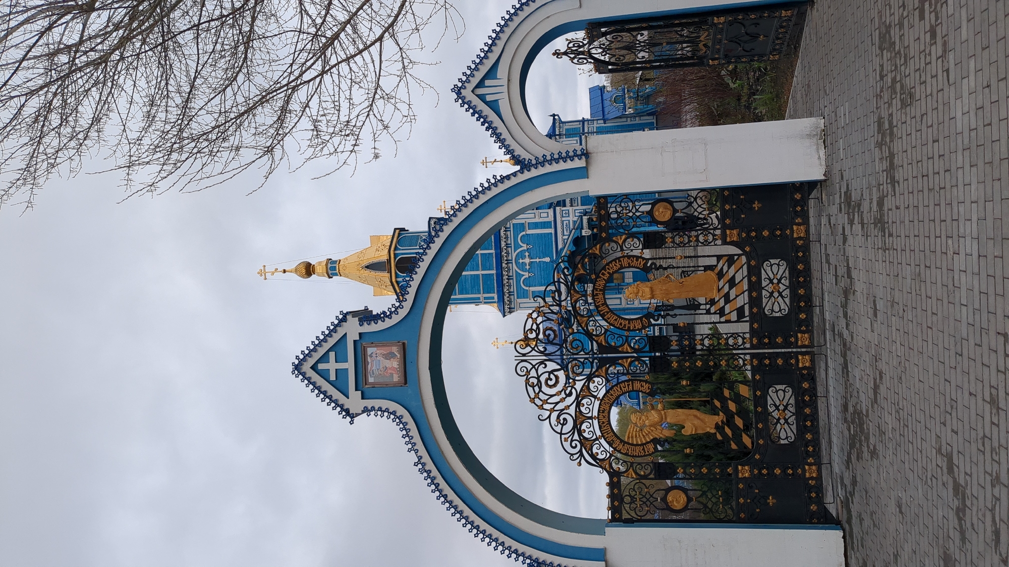 The image captures the entrance to a Russian Orthodox church in Ulyanovsk, Russia. The frame is composed of a partially visible tree on the left, the ornate entrance arch, a view of the church itself, the decorative gates, and a paved pathway on the right. The entrance arch is painted blue and white with intricate detailing. Through the arch, parts of the blue and white church building with a golden dome can be seen. The black and gold gates are ornamented with golden figures. The sky is overcast and grey. The pavement is made of grey rectangular blocks. The photo is taken from a ground-level perspective.