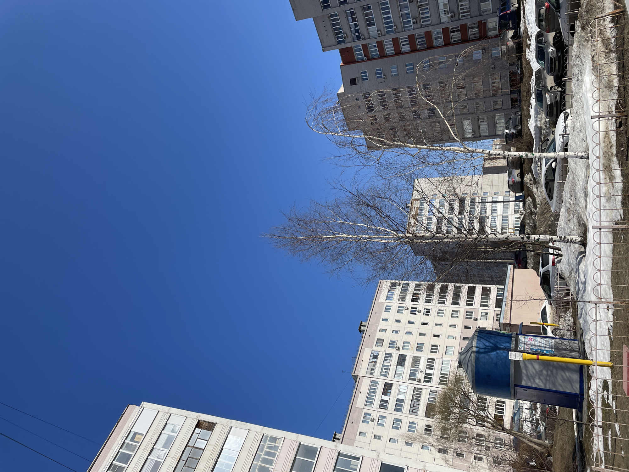 The image captures a street scene in Izhevsk, Russia, showcasing a view of residential buildings against a clear blue sky.
**Environment:** The scene is bathed in daylight, suggesting a bright, sunny day. Patches of melting snow indicate it is likely early spring.
**Buildings:** Multiple Soviet-era apartment blocks surround the frame. These buildings feature rectangular architecture and repetitive window patterns.
**Vegetation:** Bare trees, devoid of foliage, frame the buildings, suggesting early spring before the onset of greenery.
**Objects:** A blue and grey structure sits on the sidewalk, likely a small kiosk or vending structure.
**Other Details:** There are cars parked along the side of the road with traces of snow. The image is taken from a low-angle perspective, looking upward toward the sky.
