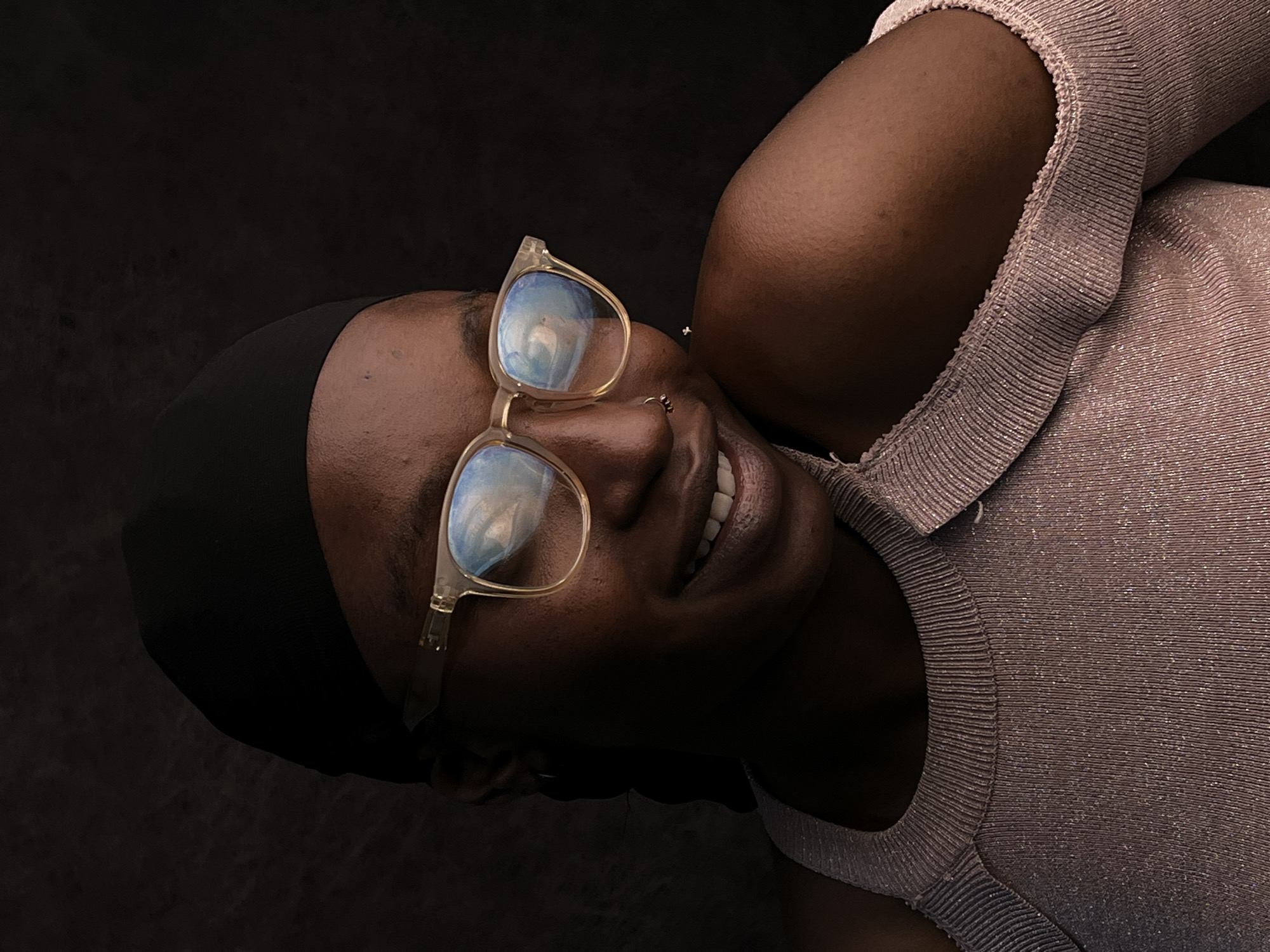 The image shows a dark-skinned woman wearing a black head covering and eyeglasses with a clear frame. She is wearing a light-colored, textured top. She is smiling, and her teeth are visible. The background is dark and indistinct. There is no indication of the city of Barakin Akawo, Nigeria in the image.