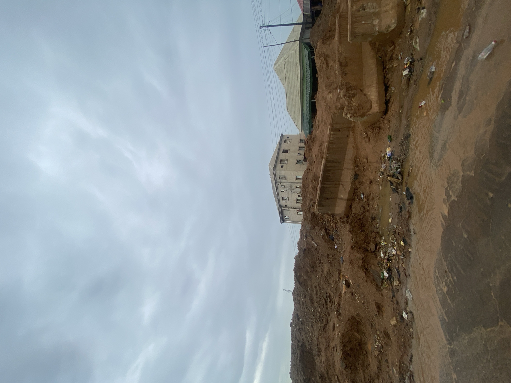 The image depicts a scene in Keffi, Nigeria, characterized by a construction site or area undergoing significant earthworks. In the foreground, piles of reddish-brown soil dominate, interspersed with scattered debris including plastic bottles, bags, and other refuse. Puddles of muddy water suggest recent rainfall or poor drainage. Concrete barriers, some appearing damaged or displaced, are visible, further indicating construction or demolition activity.
In the middle ground, two buildings are present. To the left, a multi-story structure with a light-colored facade and visible windows stands. To the right, a building with a pitched, light-colored roof and green awnings is situated. Electrical wires stretch across the scene, supported by utility poles, a common feature in Nigerian urban environments.
The sky is overcast with a uniform layer of clouds, contributing to a muted and somewhat somber lighting. The overall impression is one of an active but untidy construction zone within an urban setting in Keffi. No people are visible in the image.