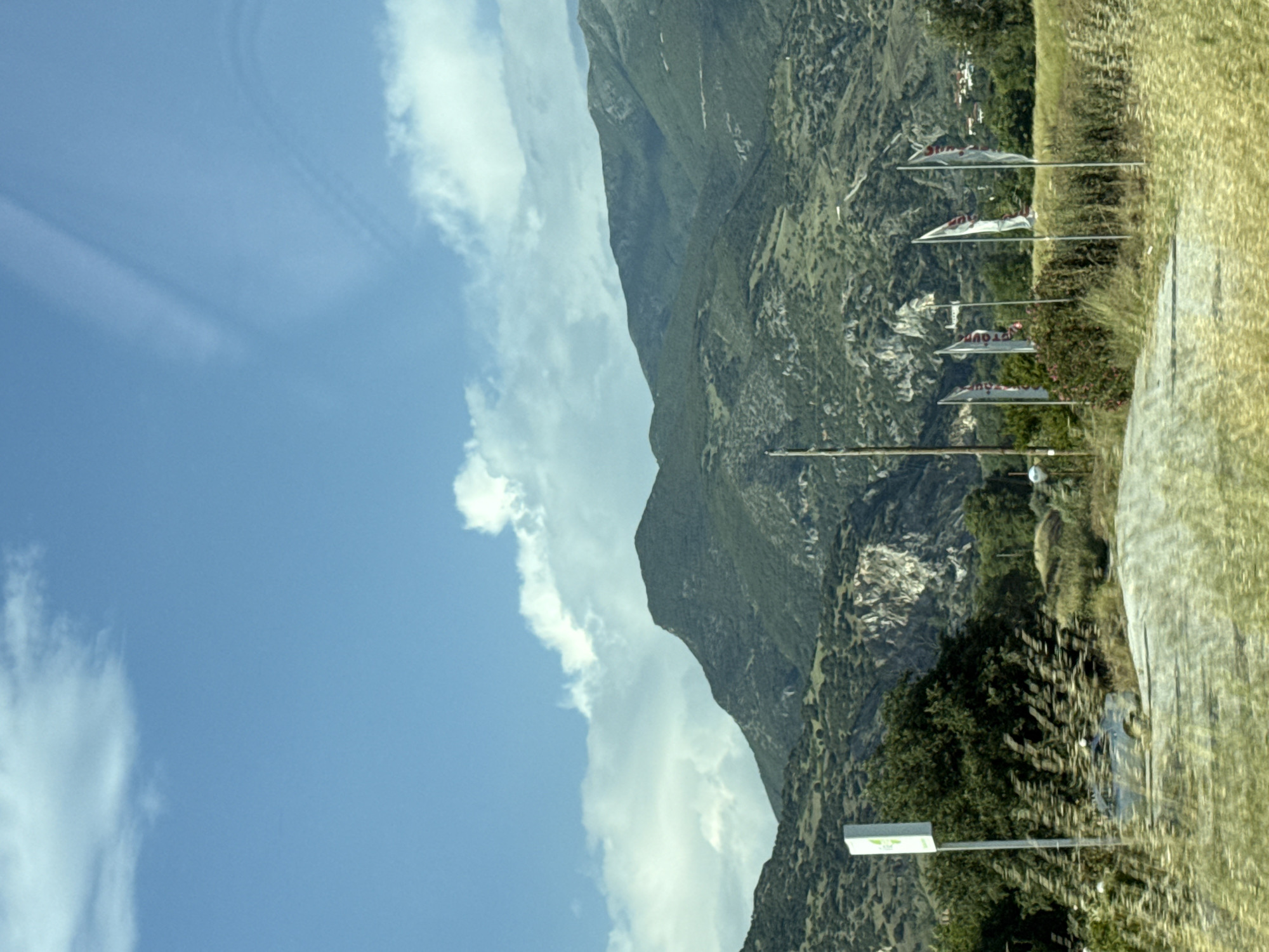 The image presents a landscape featuring a mountain range partially obscured by clouds in a blue sky. Along the roadside, several banners are visible, attached to poles. A road and adjacent grassy area are also present. A road sign is seen. Based on the banners indicating "Hot Tours", the location can be presumed to be Serres, Greece, given the context.