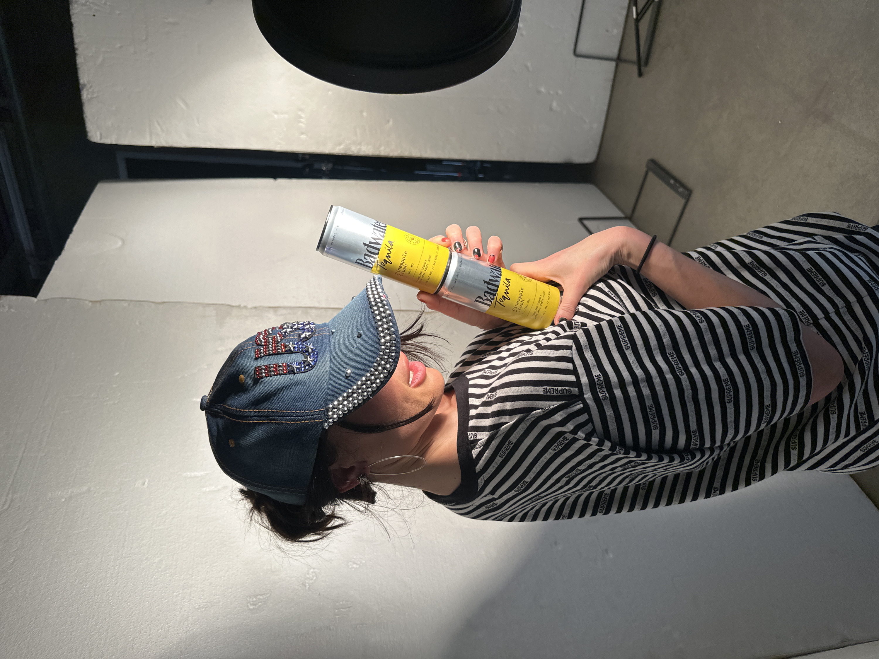 The image displays a close-up, portrait-oriented shot of a person, rotated 90 degrees clockwise, against a simple, light-colored background that appears to be part of an indoor studio or set. The lighting is bright, illuminating the subject from above and slightly to the left.
The person, seemingly a young adult, is looking slightly upwards and to their right, with their mouth slightly open, revealing pink lips. They have dark hair, some of which is styled messily and peeking out from under a cap. Their eyes are not fully visible due to the angle and possibly makeup, but dark eyeliner is apparent on the upper lash line. They are wearing large, thin hoop earrings.
They are dressed in a short-sleeved, black and white striped top. Upon closer inspection, the black stripes contain repeated white text that reads "SUPREME". The person's left arm is visible, bent at the elbow, and they are holding two slender, silver-colored cans in their left hand, stacked one on top of the other. The cans have a bright yellow label with "Badwater Tequila" prominently displayed. Further text on the label includes "Pineapple Crush", "SPARKLING TEQUILA WITH REAL FRUIT JUICE", "ALL NATURAL", and details about alcohol volume and calories ("% ALC VOL // 363 KCAL / 200ML"). Their fingernails are painted with a black and red design. A black hair tie is visible on their wrist.
The person is also wearing a denim baseball cap. The brim of the cap is heavily embellished with small, silver-colored studs. The front of the cap features an ornate design made of red, white, and blue rhinestones, forming what appears to be the number "23" or possibly a stylized "USA" or "US" with a star design.
The background consists of plain, off-white surfaces, possibly walls or panels, creating a clean, minimalist setting. There are some faint textures and lines on these surfaces. In the upper right corner of the original image (which would be the upper background if oriented correctly), part of a dark, metallic structure, possibly a stand or furniture, is visible. Another dark, horizontal line suggests a seam or a shelf in the background. The overall impression is a focused product shot with the person as a model.