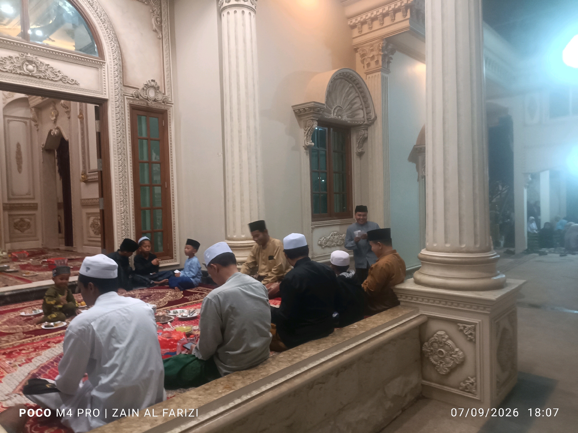 The image captures a gathering of men and boys seated on a large, ornate rug inside a building. The setting appears to be a formal or religious space, indicated by the presence of large columns with decorative bases, ornate wall carvings, and a recessed entryway. Several individuals are wearing traditional Islamic attire, including peci (caps) and long garments. Some are engaged in conversation, while others appear to be resting or observing. In the foreground, a man in a white shirt and peci faces away from the camera, and to his left, another man in a grey shirt and peci is also seated. The lighting is somewhat dim, with some bright, diffused light sources visible in the background, suggesting an indoor setting potentially during the evening. The overall atmosphere seems calm and communal. The timestamp overlay at the bottom right corner indicates the date as 07/09/2026 at 18:07. The watermark "POCO M4 PRO | ZAIN AL FARIZI" is present in the bottom left corner. While the image depicts a scene that could be common in many places, the provided context specifies the location as Buduran, Indonesia.