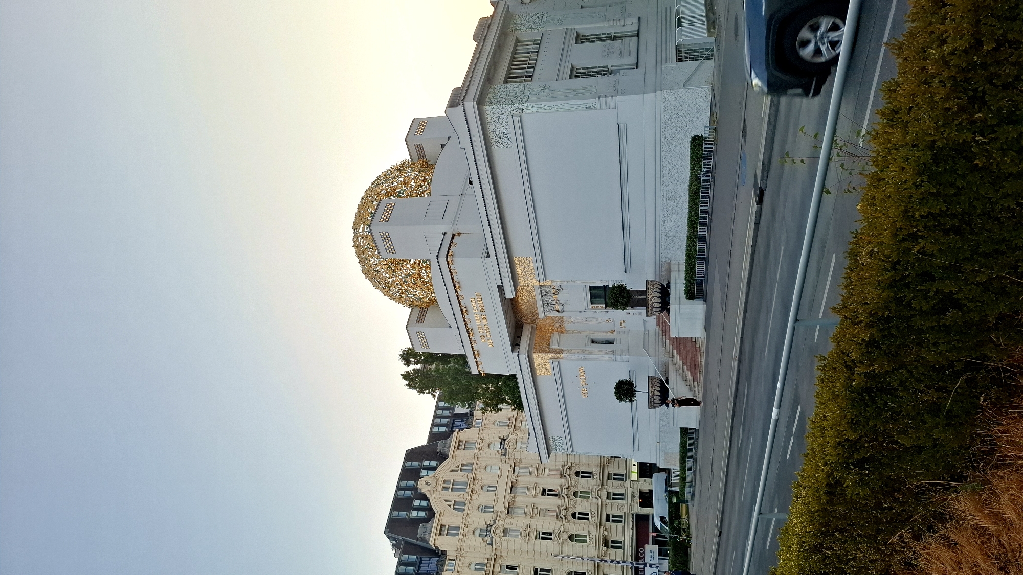The image captures the Secession Building, a prominent landmark in Vienna, Austria. The building, characterized by its white facade and ornate golden dome, stands out against a clear, pale blue sky. The dome, a signature feature, is covered in gilded leaves. In front of the Secession Building, a set of stairs leads up to an entrance, flanked by potted trees.
To the left of the Secession Building, an imposing, multi-story residential building with a mansard roof and numerous windows is visible. This building exhibits a more traditional architectural style, contrasting with the Secessionist design.
In the foreground, a street runs horizontally across the image, with a light gray railing bordering a landscaped area. The landscaping consists of manicured green hedges and some dry grasses. A dark-colored SUV is captured in motion on the street, its front-right wheel and part of the body visible as it drives away from the camera. A person in dark clothing is standing on the stairs of the Secession Building, facing towards the entrance. The overall scene suggests a late afternoon or early evening setting, with soft lighting.