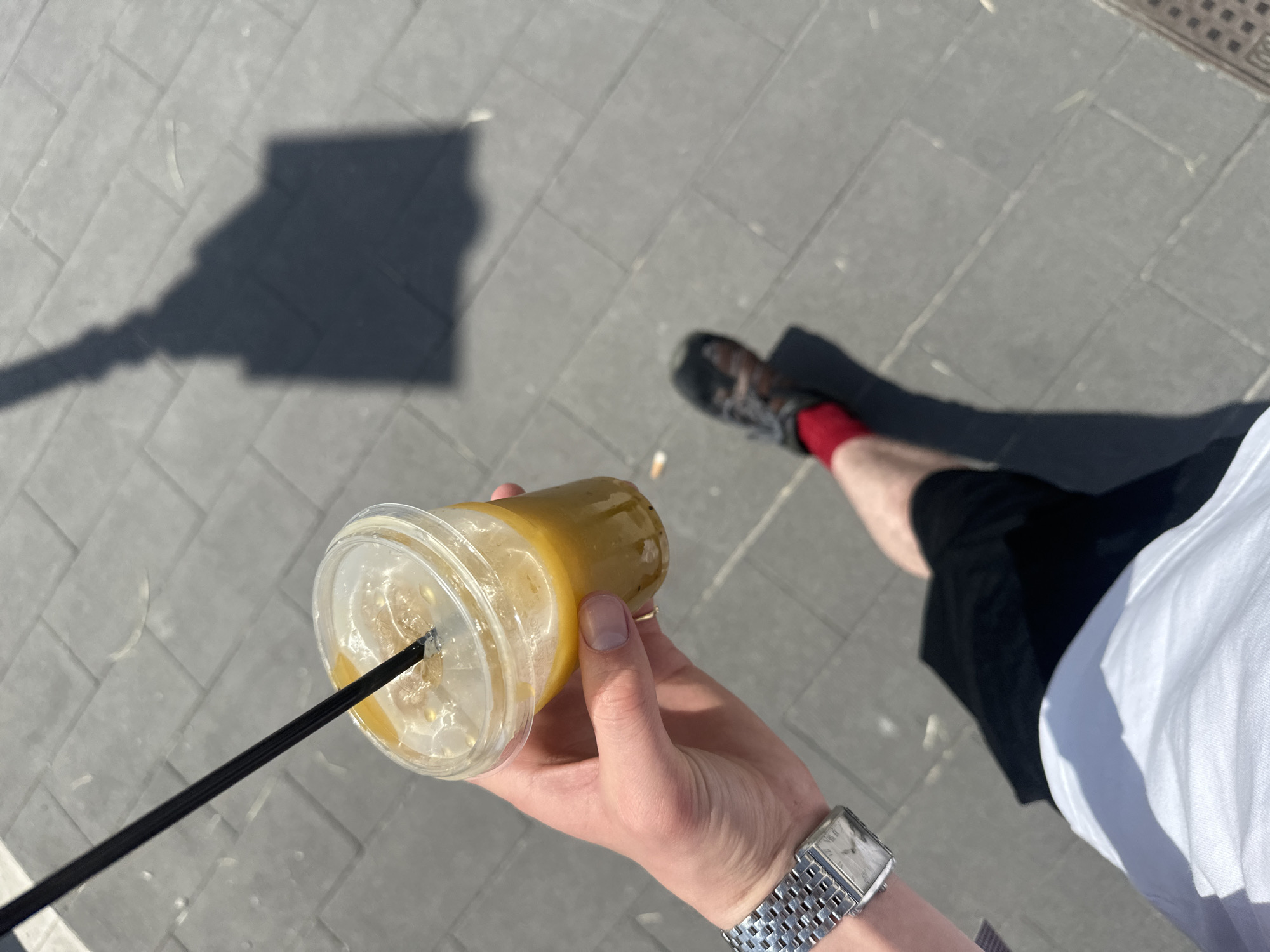 The image presents a first-person, high-angle perspective of a person walking or standing outdoors on a sunny day, likely in Lipari, Italy. The main subject is a hand, positioned in the lower center, holding a clear plastic cup with a yellowish-orange liquid, possibly a fruit smoothie or juice, containing ice or pulp. A black straw protrudes from the cup's lid. A metallic watch with a rectangular face and a link bracelet is visible on the wrist of the hand holding the cup.
In the mid-ground, the person's leg is visible, clad in a dark shoe with laces and a vibrant red sock, along with a segment of dark shorts or trousers and a white t-shirt. The ground is paved with rectangular gray tiles. Strong, well-defined shadows are cast across the pavement, indicating bright sunlight and suggesting it is midday or early afternoon. A prominent, elongated shadow of what appears to be a lamppost or a similar vertical structure stretches across the upper left quadrant of the frame. The shadow of the person holding the drink is also visible on the ground. No visible text is present in the image.