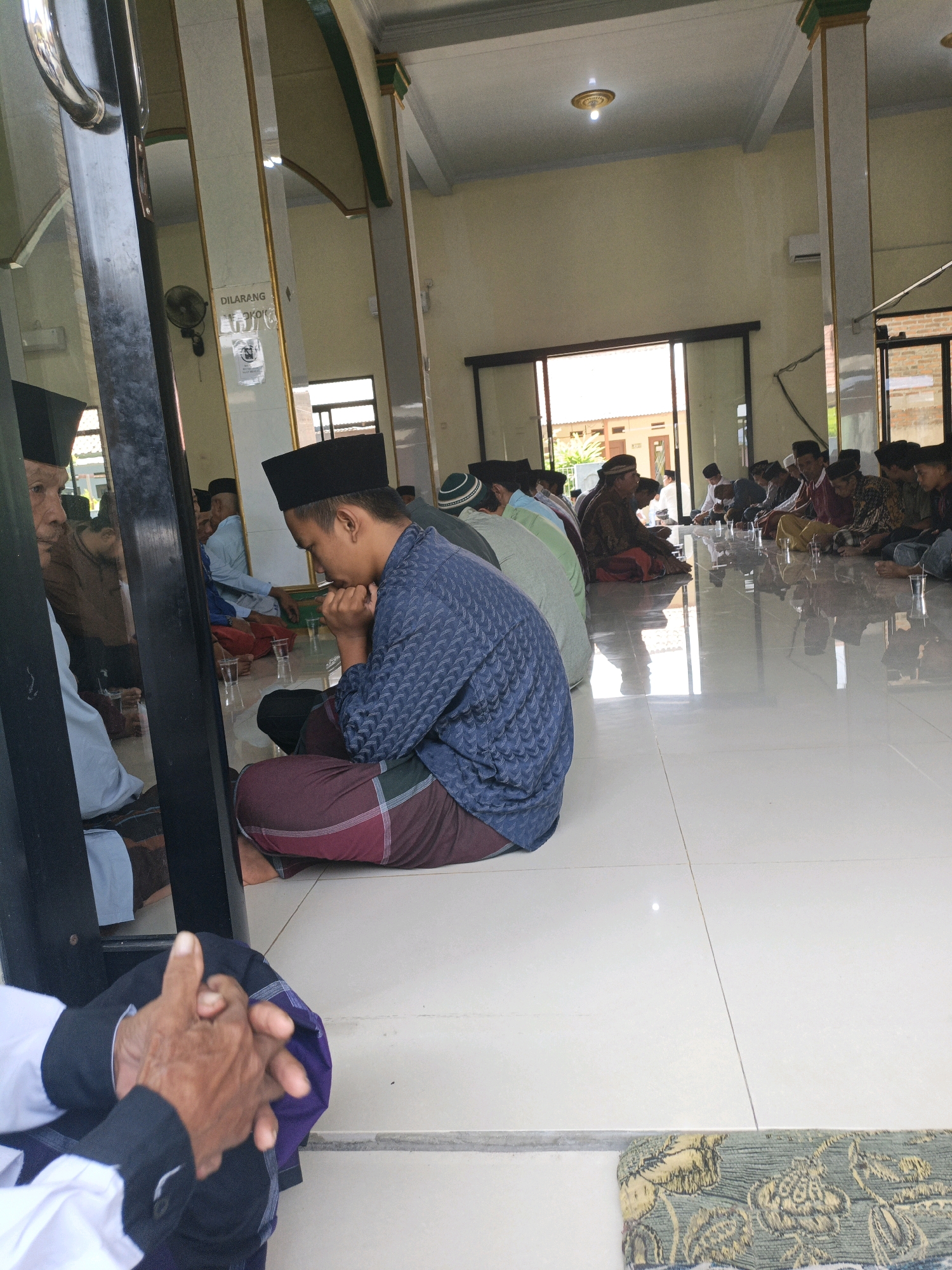 The image depicts a gathering of men seated on the floor inside a building, likely a mosque, in Cilegon, Indonesia. The men are all wearing peci hats, and many are dressed in sarongs. Some are engaged in prayer or contemplation, with their hands clasped or resting in their laps. In the foreground, two older men are visible, one with his hands interlocked, and the other partially obscured by a dark pillar. The room has a tiled floor that reflects the light from the ceiling and the open doorways. Beyond the seated men, the space opens to an outdoor area with buildings visible in the distance, suggesting an urban or semi-urban setting. A sign on a pillar reads "DILARANG" which translates to "FORBIDDEN" in Indonesian, indicating rules or restrictions within the area. The overall atmosphere appears to be one of solemnity and community.