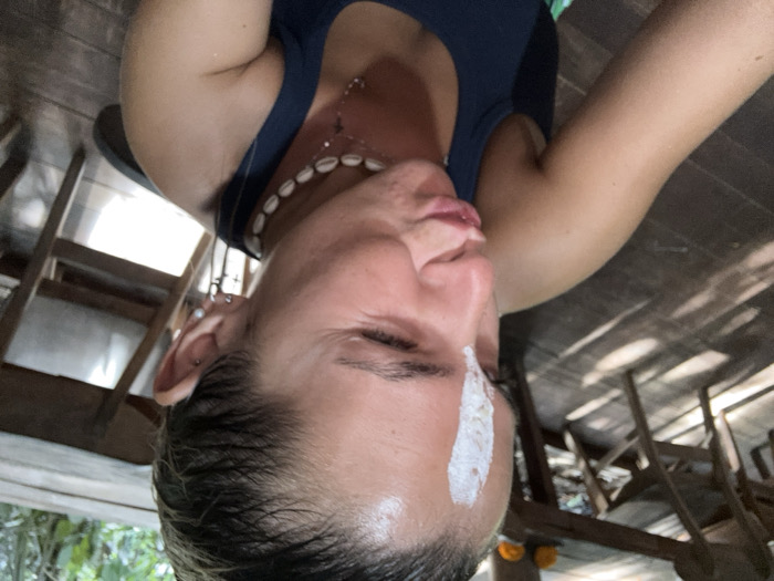 An individual is captured in a close-up, selfie-style photograph within a semi-open wooden structure in Punggul, Indonesia. The person, appearing to be female, has slicked-back dark hair and fair skin. A distinct vertical stripe of white substance is applied centrally on the forehead. The individual's right eye is closed in a wink, and the lips are puckered. She wears a dark-colored tank top or sports bra. Accessories include multiple earrings, one appearing as a cross charm, a white shell choker necklace, and a delicate silver-colored chain necklace with a small cross pendant. The background features a blurred view of a dark wooden plank floor with prominent shadows, along with partial views of wooden tables and chairs. Two small orange objects are visible on a distant table. Green foliage is discernible in the upper right background, indicating proximity to natural vegetation.