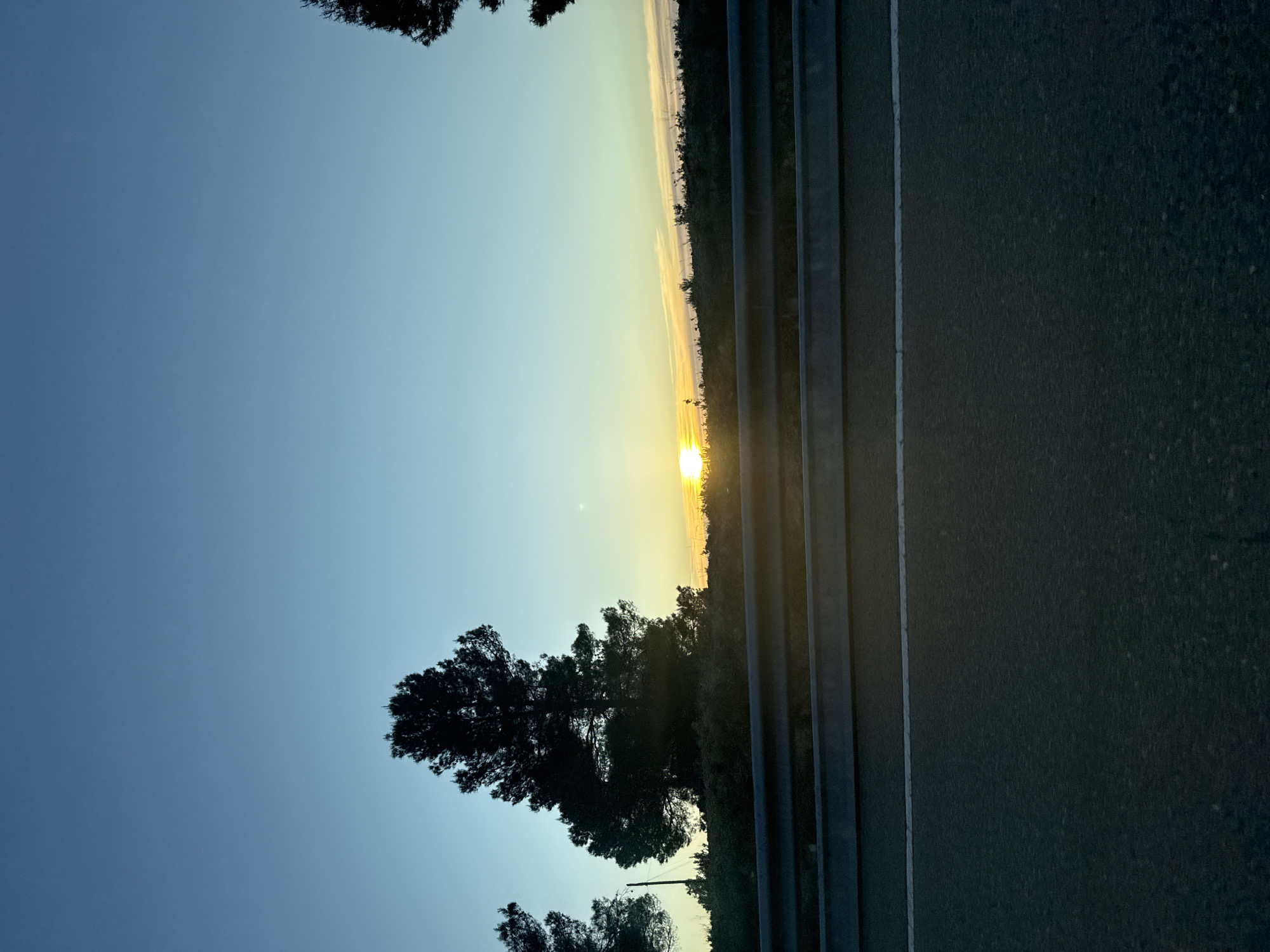 The image shows a sunset viewed from what appears to be a roadside location. The sky is a gradient of blue to yellow-orange, indicating the time of day. A bright sun is visible on the horizon. Silhouetted trees and vegetation appear against the bright sky. To the right of the vegetation, a road or paved surface is seen, with visible lines. The location appears to be Fórnoles / Fórnols, Spain based on the presence of Mediterranean vegetation. There are no people visible in the scene.