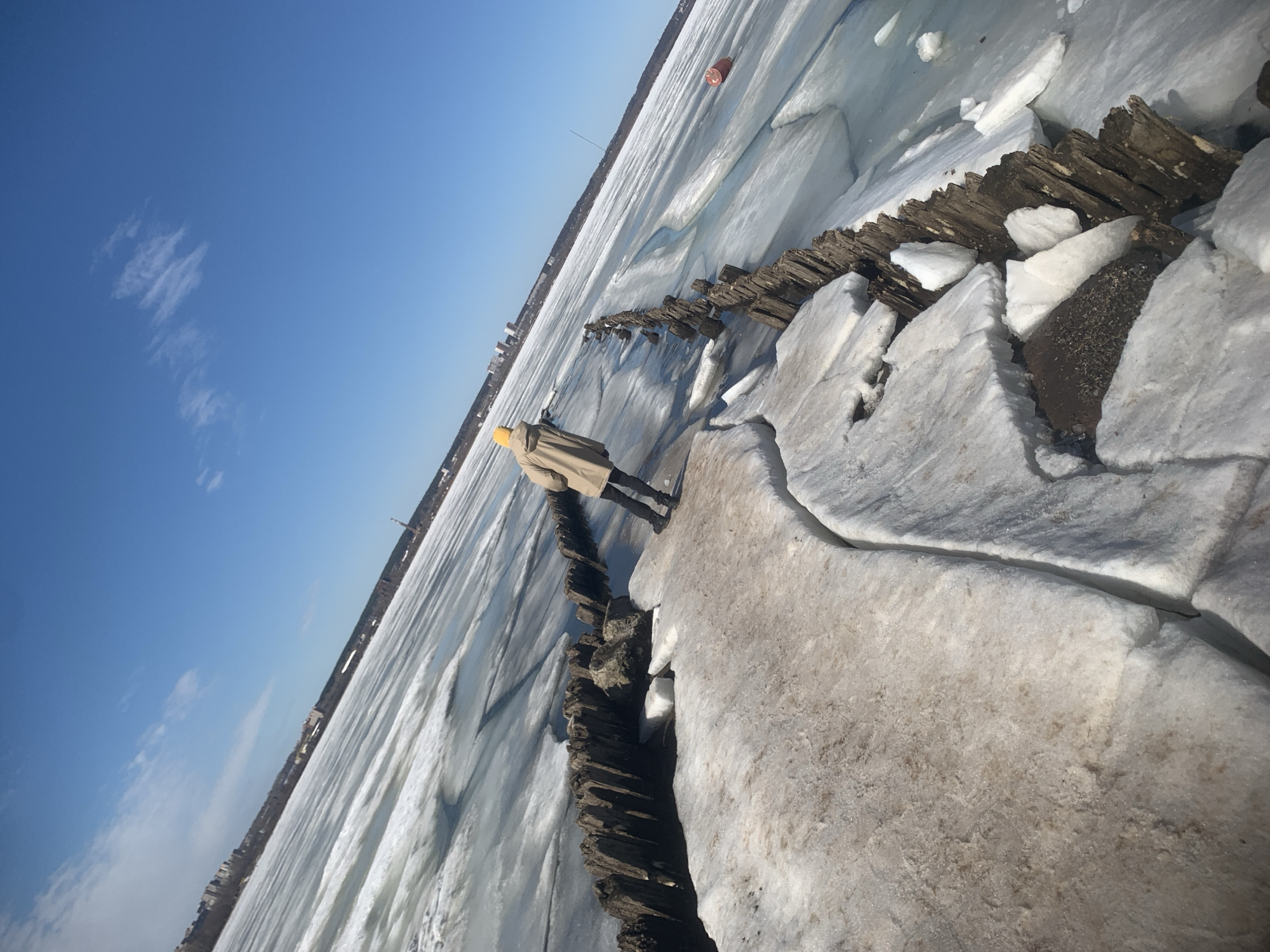 A person in a tan coat and yellow hat stands on a wooden structure, seemingly a breakwater, along a frozen body of water, likely a river or lake, in Izhevsk, Russia. The landscape is dominated by ice and snow. The sky is blue with a few clouds. In the distance, the city skyline is visible.