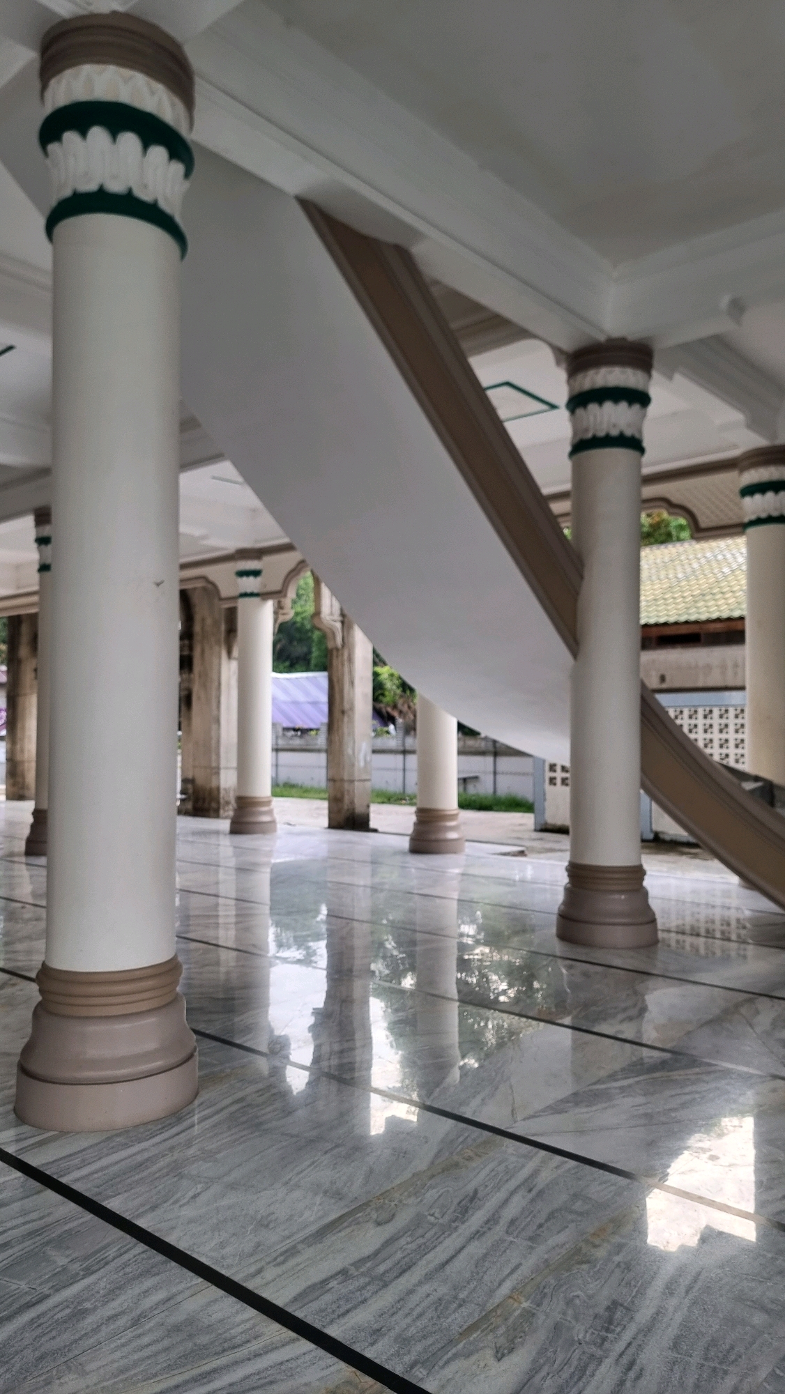 This image depicts the interior of a mosque in Lhokseumawe, Indonesia, focusing on the architectural features of the building. The foreground is dominated by a polished marble floor with a grey and white veined pattern, reflecting the overhead structure. Several large, white cylindrical columns with decorative bands of brown and green are visible. These columns support a white ceiling and a sloping architectural element, possibly a ramp or an extended staircase, which is also white with brown trim.
In the background, through the open colonnade, glimpses of the exterior environment are visible. There are other columns, some appearing older and more weathered. Beyond these, trees and foliage can be seen, along with what appears to be a building with a tiled roof. Faintly visible are structures that suggest an urban setting, possibly indicating the mosque's location within the city of Lhokseumawe. The overall impression is one of a clean, well-maintained, and serene religious space. There are no people or animals present in the image, nor any discernible actions or interactions. The lighting suggests it is daytime, with natural light filtering in.