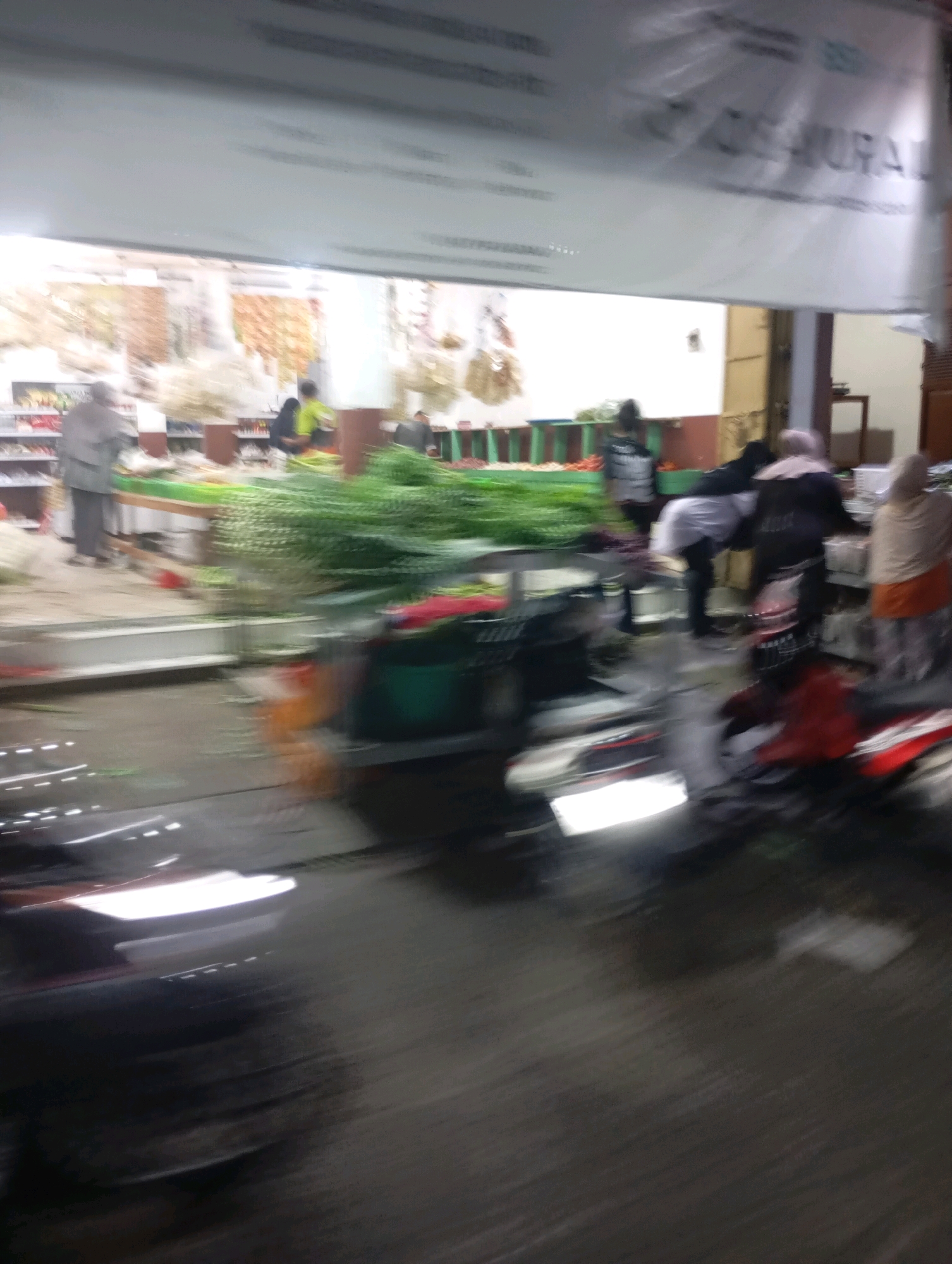 A street scene in Babelan, Indonesia, captured at night, is depicted with significant motion blur. The image appears to be taken from a moving vehicle, possibly a motorcycle, indicated by the streaks of light and blurred foreground elements. In the background, several individuals are visible, seemingly engaged in market activities. One person, wearing a grey garment, is standing near a stall laden with fresh produce. Another individual in a bright green shirt is also present, along with others in darker clothing. A large white banner with some text is stretched across the top of the frame, partially obscuring the view of the sky. The ground is wet, suggesting recent rain. The overall impression is one of a bustling nighttime market with limited visibility due to the blur.