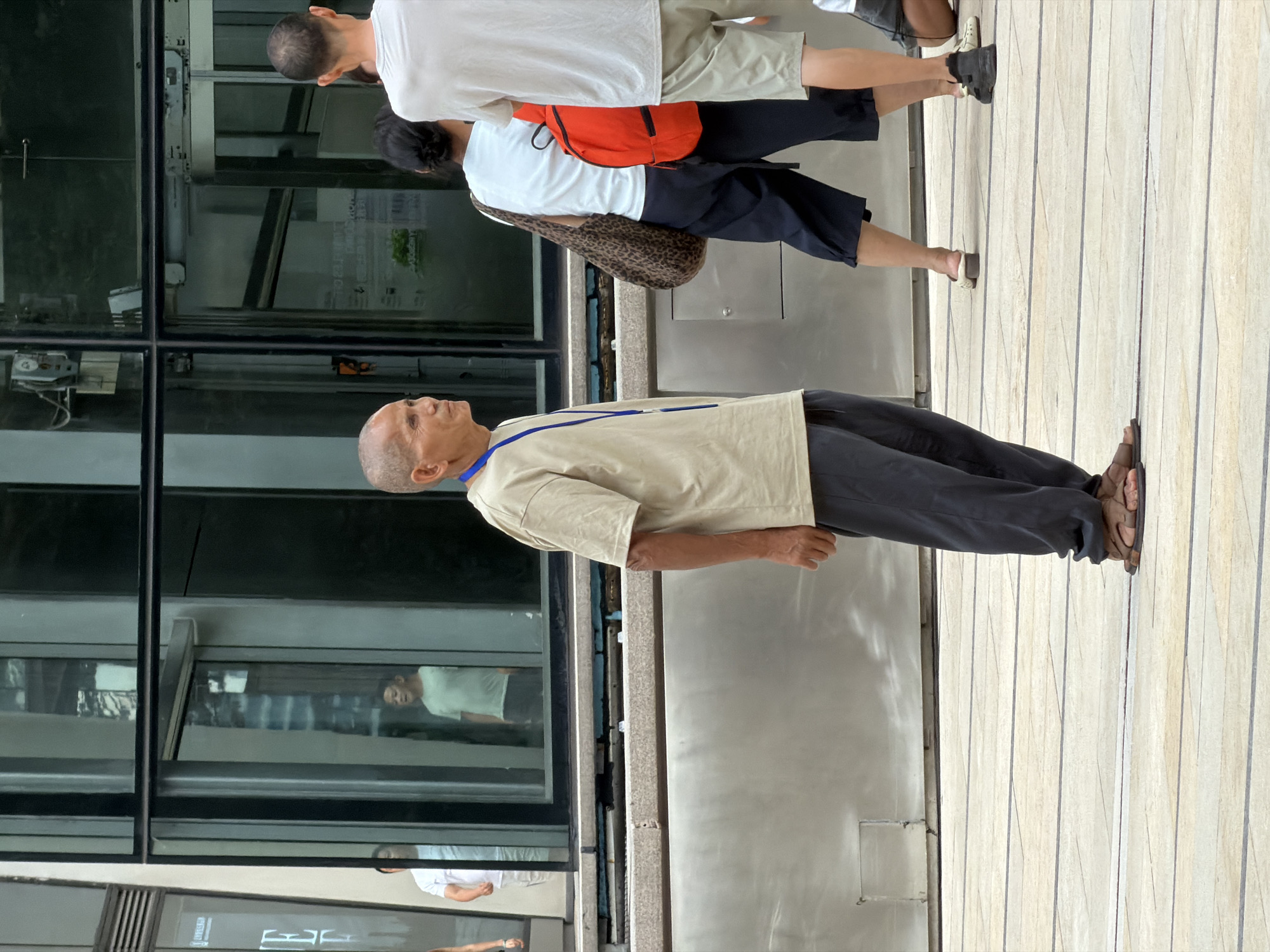 In Yuzhong District, China, an elderly East Asian man stands in the foreground facing left, wearing a beige t-shirt and dark trousers. He has a bald head and a blue lanyard around his neck. He is wearing brown sandals. Behind him, a woman with dark hair wears a white t-shirt, dark capri pants, and carries a leopard-print bag over her left shoulder and a bright orange backpack on her back. To the right of the woman, a person in a white t-shirt and light-colored shorts stands facing away from the camera. In the background, through large glass windows of a modern building, several individuals are visible, including a man in a light green t-shirt and dark shorts. The scene appears to be an outdoor public space with a tiled ground surface.