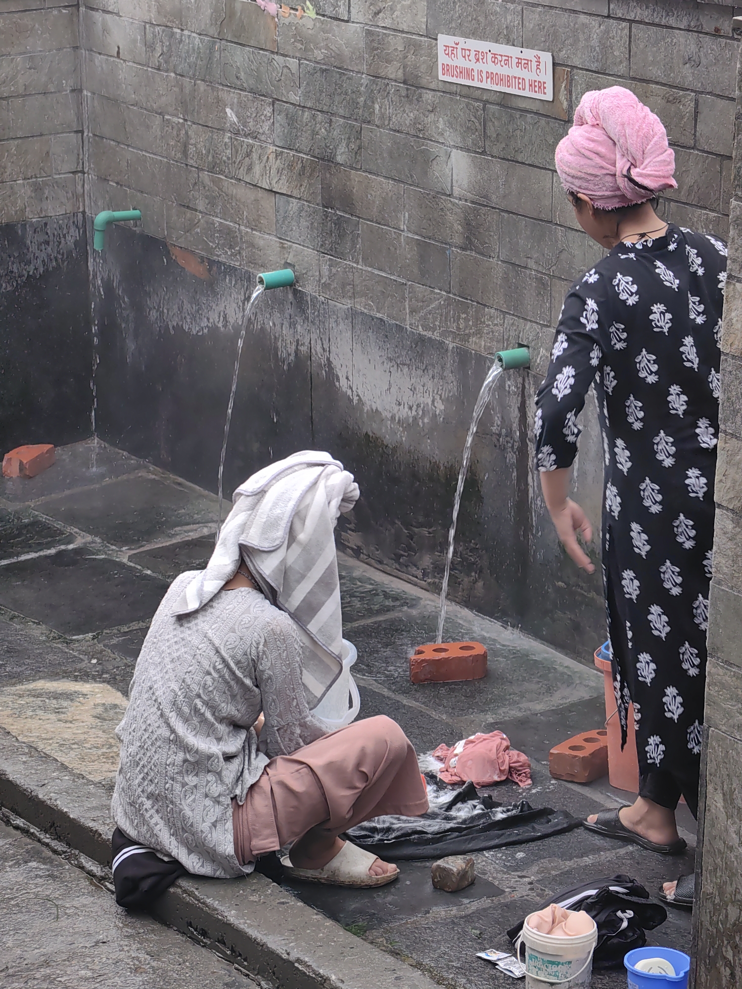 In Vashisht, India, two individuals are seen by a public washing area. On the left, a person wearing a grey patterned top and pink pants, with a striped towel wrapped around their head, is seated on the ground, facing away from the camera. They are positioned near a green pipe from which water is flowing. On the right, another individual, wearing a black and white floral patterned garment and a pink headscarf, stands and reaches towards a green pipe where water is also flowing. In the foreground, several items are scattered on the wet ground, including bricks, clothing, buckets, and footwear. The wall behind the subjects is constructed of grey stone blocks, and a red and white sign in Hindi and English, reading "BRUSHING IS PROHIBITED HERE", is affixed to the wall.