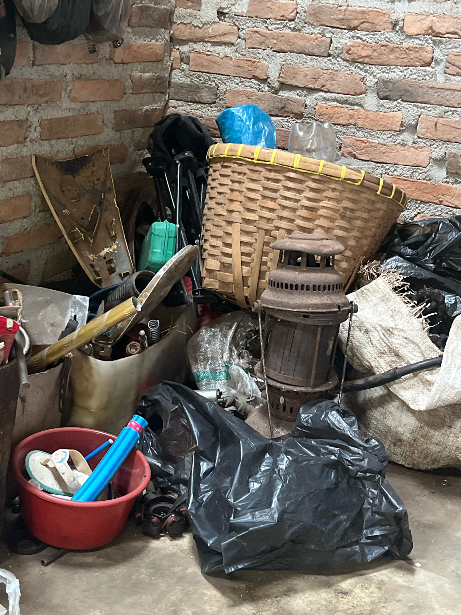 The image depicts a cluttered corner of what appears to be a storage area or shed in Kebumen, Indonesia. The walls are constructed of rustic red brick with visible mortar. Various items are piled haphazardly, suggesting disarray. Dominating the scene is a large, woven basket filled with other objects, including a blue plastic bag and a crumpled plastic bottle. In front of the basket sits a rusted, old-fashioned oil lantern. To the left of the lantern, a shovel with a golden handle rests against a pile of items, including a green plastic container. A red bucket in the foreground contains a pair of flip-flops and a blue tube. Black plastic trash bags are scattered throughout the lower portion of the image, some appearing full. A rough, woven sack is visible to the right of the lantern, and a dark hose or pipe runs across it. The floor is concrete and appears dusty and worn. No people are visible in the image. The overall impression is one of neglected storage.