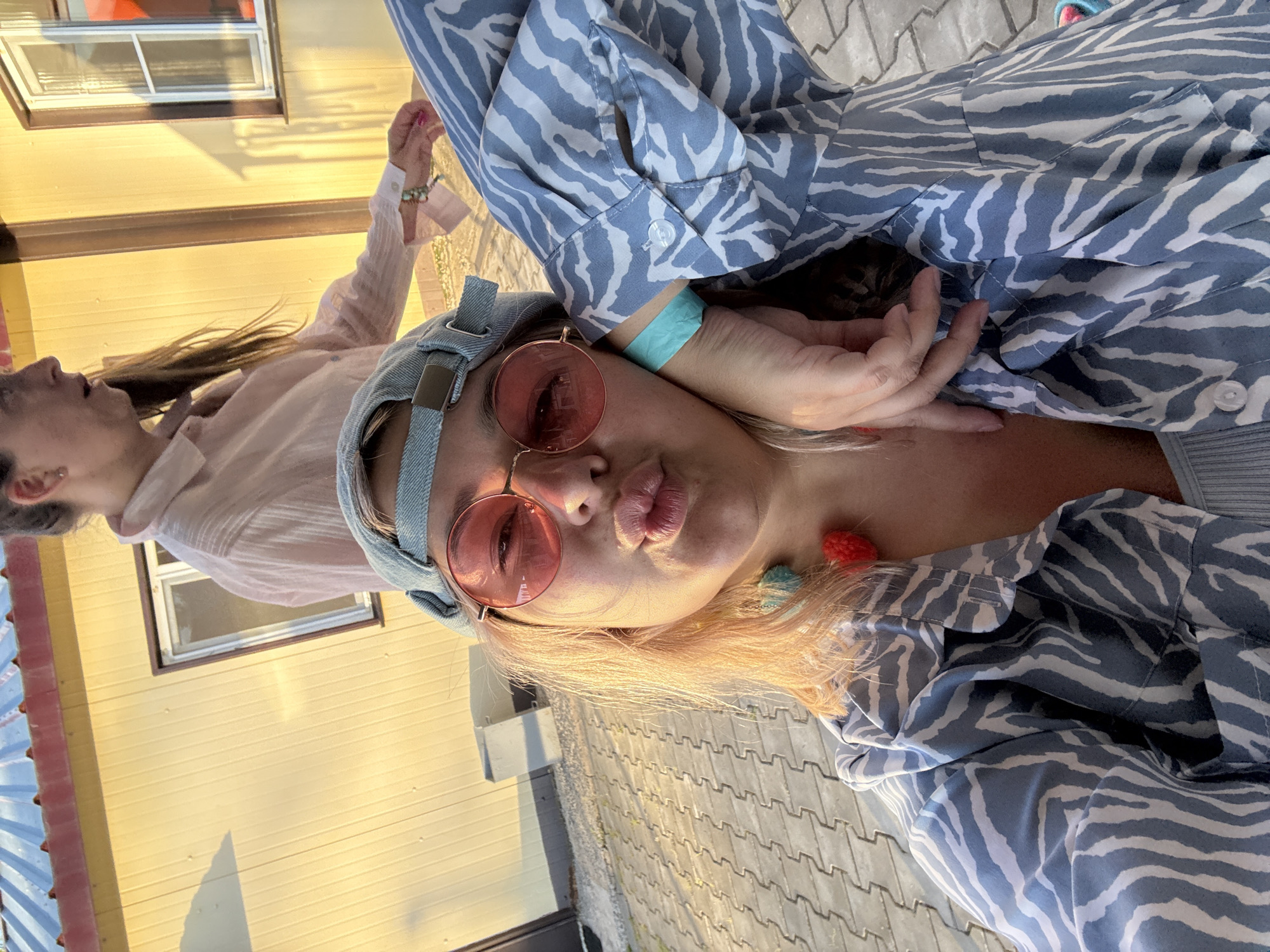 In an outdoor setting with a light yellow building with a red-trimmed roof and white-trimmed windows in the background, a woman with blonde hair, wearing a denim baseball cap, round pink-tinted sunglasses, a blue and white zebra-print shirt, and a light blue wristband, puckers her lips for a selfie. A second person, partially visible, wears a light pink collared shirt. The ground is paved with grey interlocking bricks. The lighting suggests it is either early morning or late afternoon. The presence of a wristband may indicate a paid event or access pass. No specific landmarks of Birobidzhan, Russia are visible in the frame.