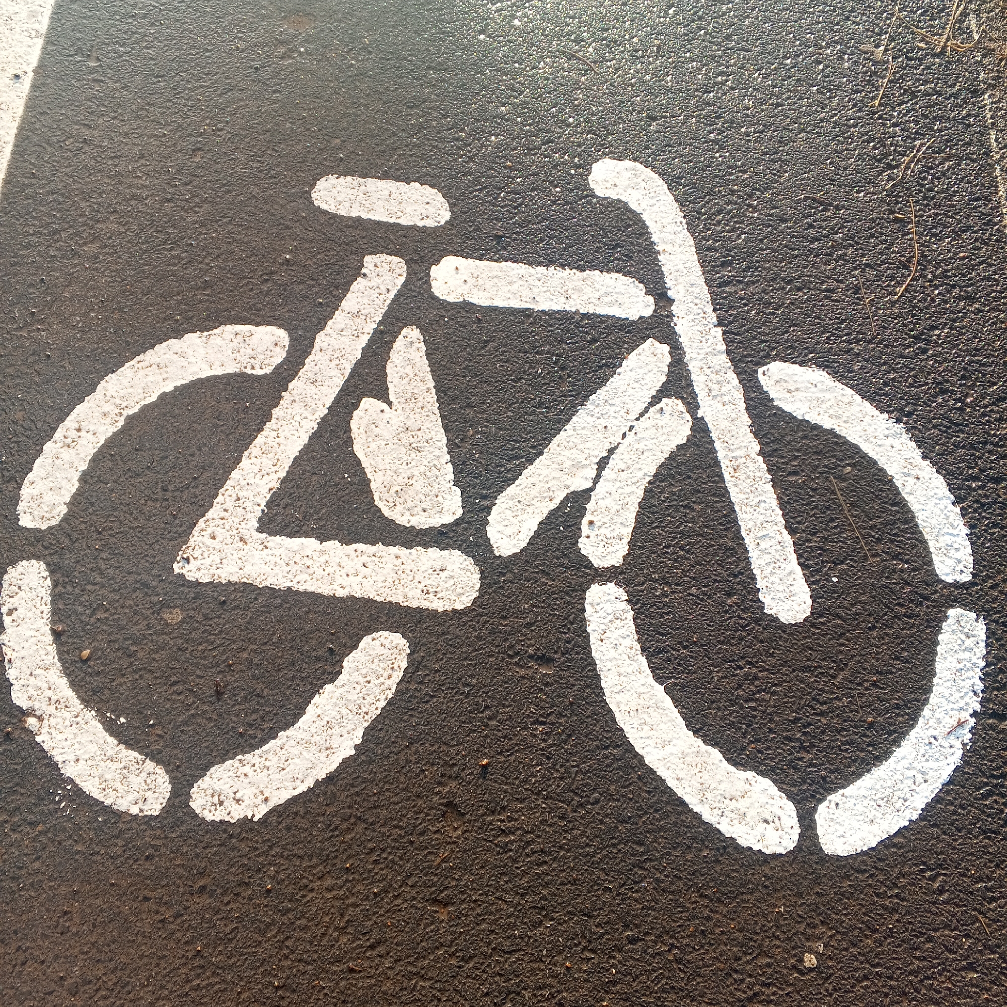 A white stencil of a bicycle is painted on an asphalt surface. The bicycle is depicted from a side view, with two wheels, a frame, handlebars, and pedals. A white line runs along the left edge of the image, parallel to the bicycle stencil. The asphalt appears textured and dark, with some small pebbles and debris visible. The scene is lit from above, casting a slight shadow. The image is cropped closely to the stencil, with no other objects or people visible. The location is specified as a street in Dyurtyuli, Russia.