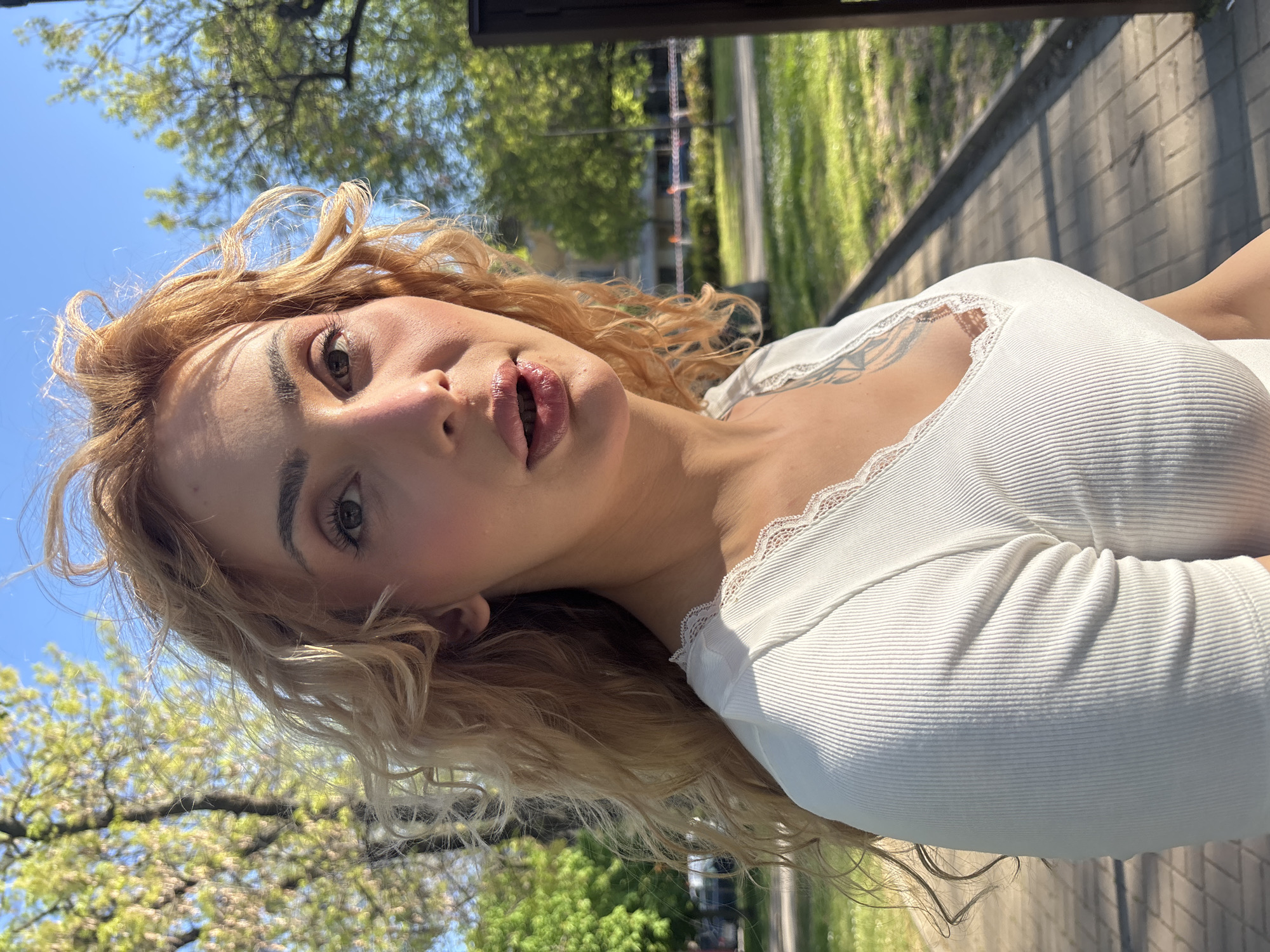 A young woman with long, wavy, blonde hair is photographed outdoors in Sofia, Bulgaria, against a backdrop of green trees and a blue sky. She wears a white, long-sleeved top with lace trim. A glimpse of a tattoo is visible on her upper chest. The background includes elements suggestive of a park or green space, with trees and a sidewalk.