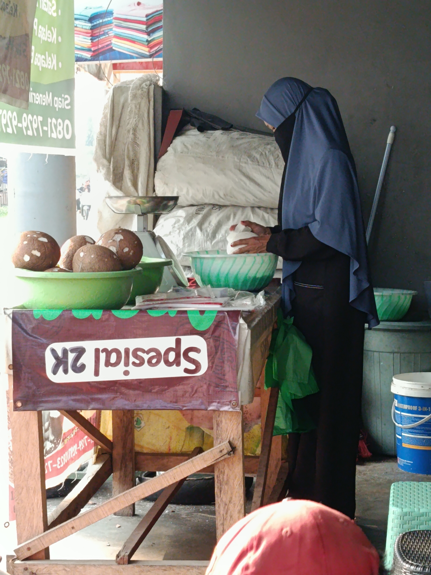 A full shot from a low angle captures an outdoor market stall, likely in Palembang, Indonesia, due to the presence of "spesial 2k" signage, a local term. A woman wearing a blue hijab and a black niqab is seen from the side, reaching into a large green bowl filled with coconuts. The stall is adorned with a brown banner featuring the "spesial 2k" logo and green accents. Behind the woman, a scale is visible, along with stacks of white, bagged goods and a light-colored textile hanging. A bright green plastic container sits to the right, near a blue bucket labeled "WATERPROOF 3-IN-1." The background reveals more market elements, including stacked clothing in vibrant colors and what appears to be other stalls. The lighting suggests it is daytime, with sunlight casting shadows.