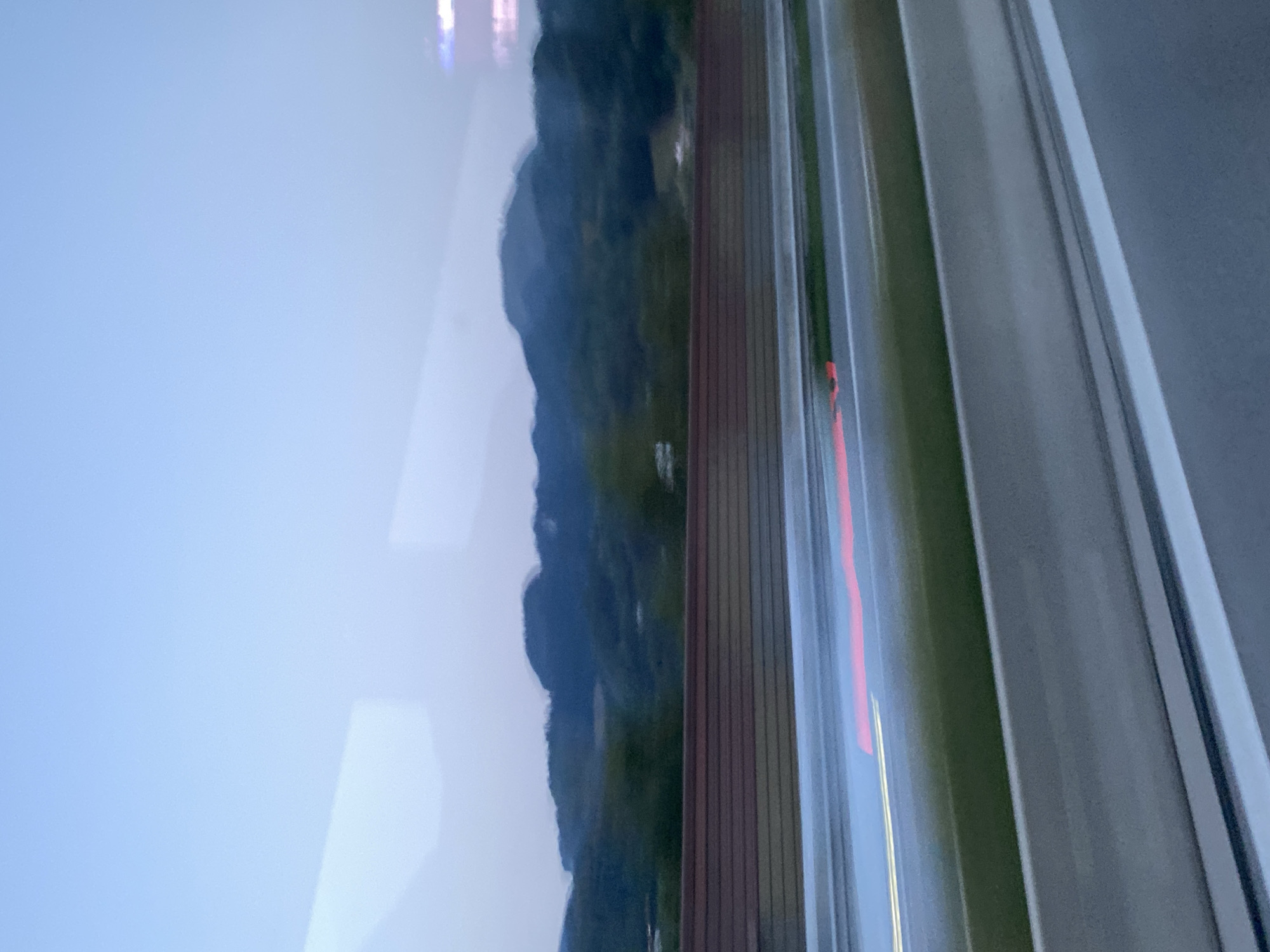 The image captures a blurred scene from a moving vehicle, likely on a highway, in Oberaudorf, Germany. The sky is a pale blue, transitioning to a darker hue towards the horizon. In the distance, blurred green hills and mountains rise against the sky. In the foreground, a highway is visible with light trails from other vehicles indicating motion. A red taillight streak is prominent across the lanes, suggesting movement. A reddish-brown barrier runs horizontally across the middle ground, separating the highway from the distant landscape. The overall impression is one of speed and a fleeting glimpse of the natural environment surrounding Oberaudorf.