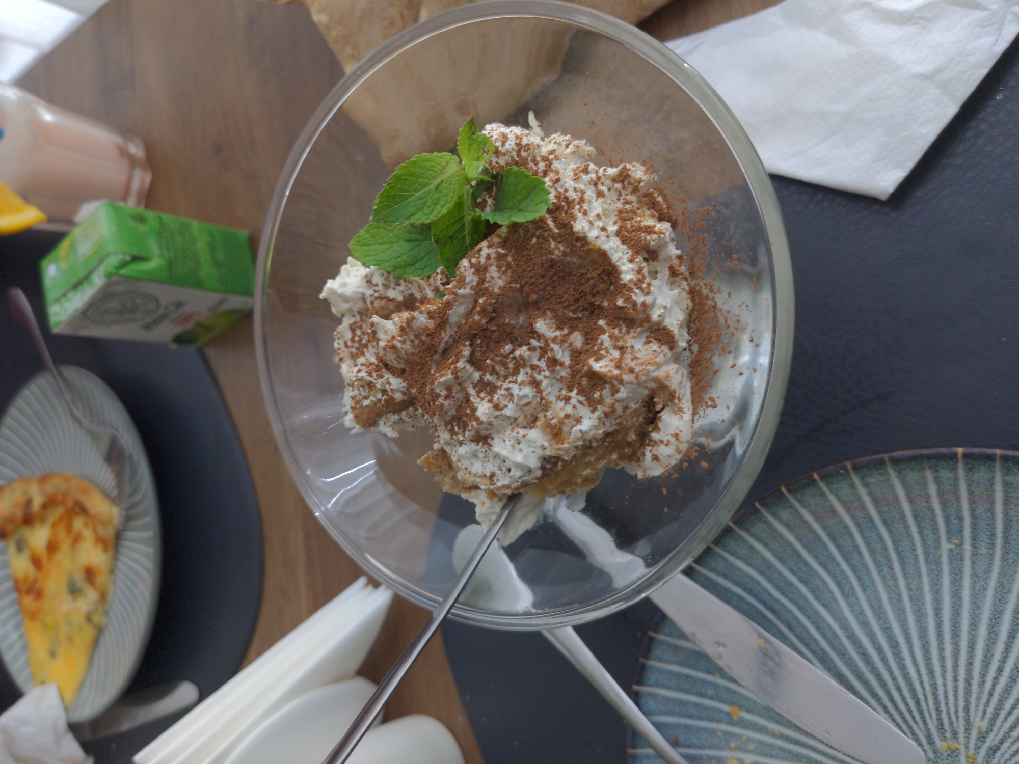 The image shows a close-up of a dessert in a clear glass bowl on a wooden table. The dessert appears to be a layered cake or pastry topped with whipped cream, sprinkled with cocoa powder, and garnished with fresh mint leaves. A fork is inserted into the dessert, suggesting it is being eaten. In the background, a slice of quiche or pie is visible on a plate, along with a green juice box and a glass of pink liquid with an orange slice. There are also white napkins and cutlery on the table. The overall scene suggests a meal or a break at a cafe or restaurant. This scene is not located in the city of Holubyne, Ukraine.