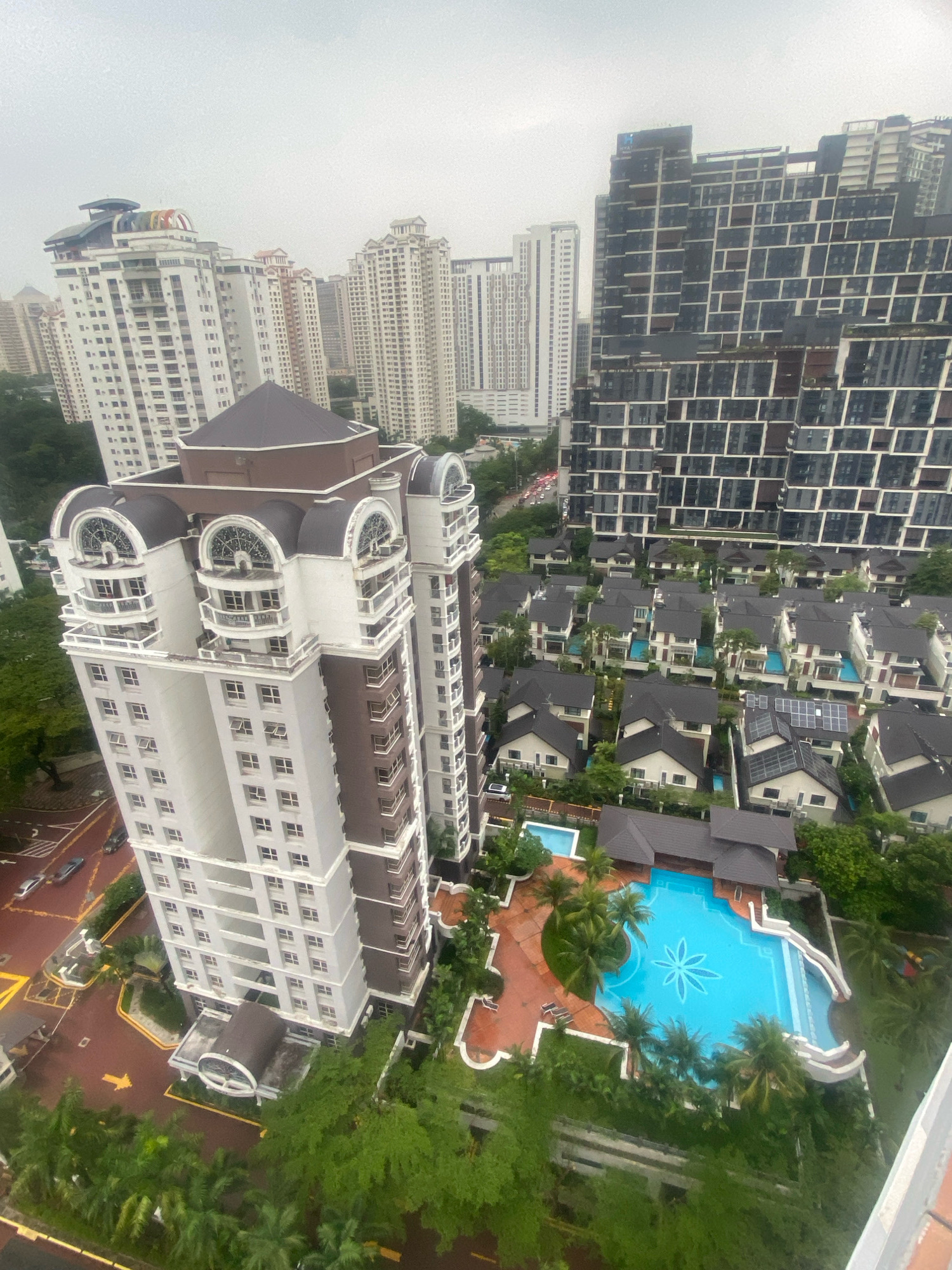 An aerial view captures a residential complex in Kuala Lumpur, Malaysia, on a cloudy day. In the foreground, a tall, modern apartment building with white and brown accents dominates the scene. It features multiple balconies, arched windows at the top, and a dark, conical roof.
Below and to the right of the apartment building, a large, ornate swimming pool with bright blue water and a white flower-like design at its center is visible. Surrounding the pool are numerous individual houses, each with dark-tiled roofs. Some of these houses appear to have their own smaller pools. A few trees and lush greenery are scattered throughout the residential area.
Further in the background, other high-rise buildings and apartment complexes of varying architectural styles can be seen, contributing to the urban landscape of Kuala Lumpur. The sky is overcast, suggesting potentially damp or rainy weather. The overall impression is one of a well-developed urban residential area with a mix of modern high-rise living and more traditional landed properties.