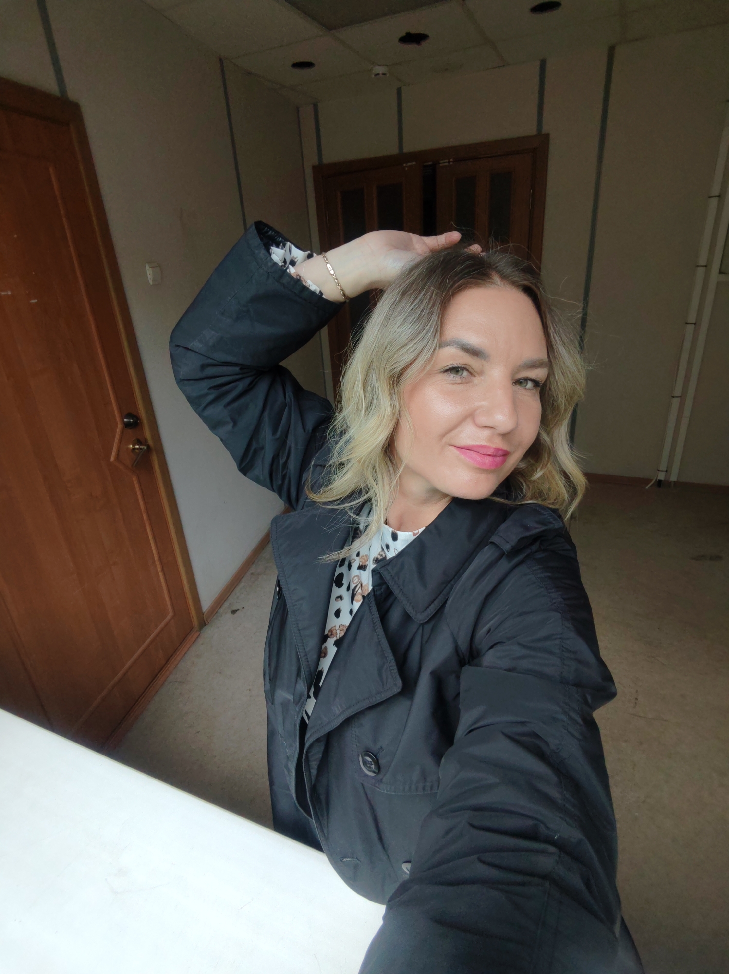 The image is a selfie of a blonde woman with light skin, pink lipstick, and green eyes. She is wearing a black jacket over a patterned shirt and has a gold bracelet on her wrist. Her right hand is raised and touching her hair.
The background appears to be an indoor space, possibly an office or apartment hallway, with wooden doors and neutral-colored walls. The ceiling has visible lighting fixtures. The location is Unknown, Unknown.