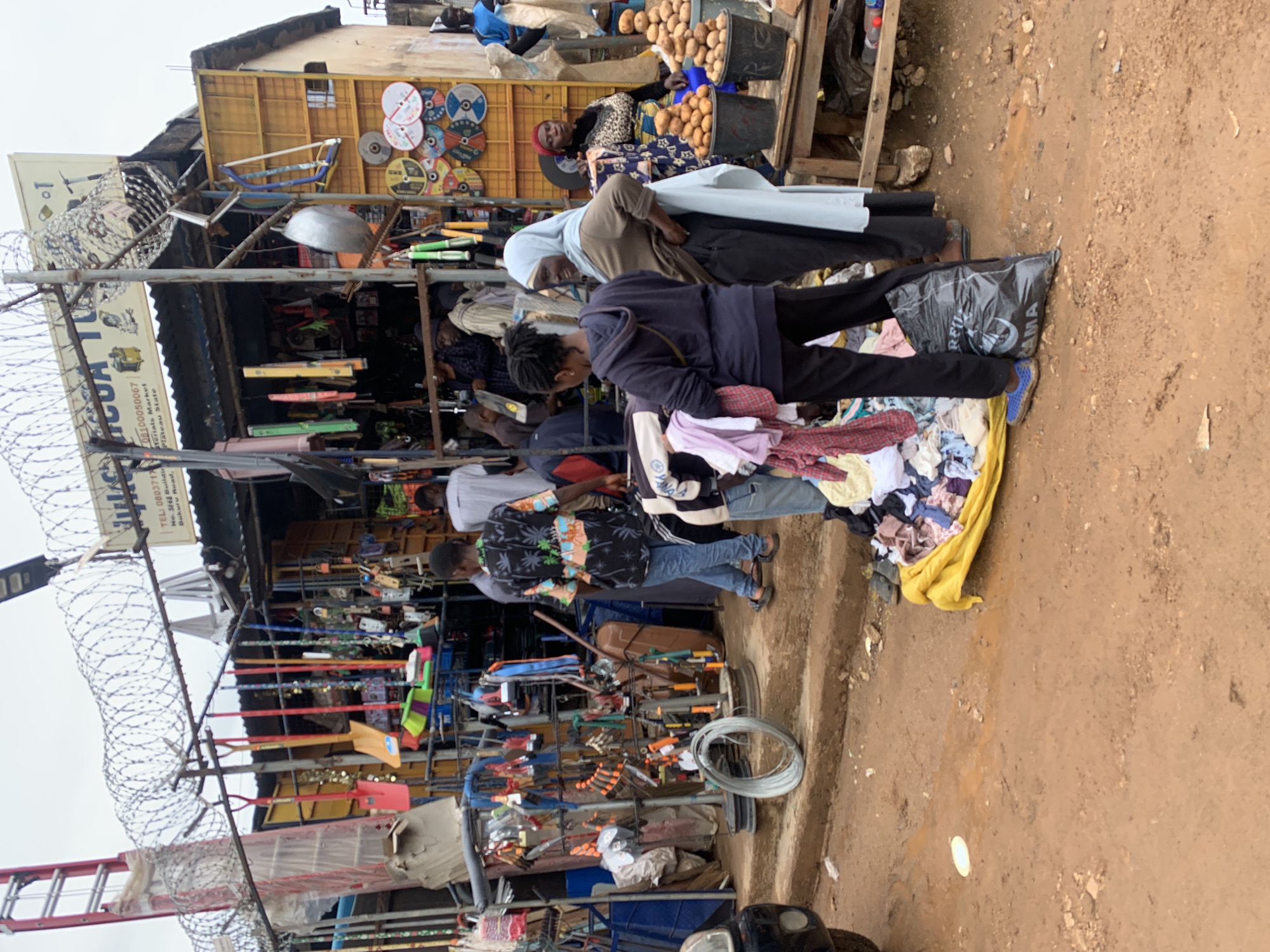 Captured at Gura Top, Nigeria, the image presents a bustling marketplace. People are visible browsing through a variety of goods. One individual is examining clothes spread on a yellow sheet on the ground. Overhead, a person wearing a hijab stands next to them. Several vendors are seen attending their respective stalls, which are densely packed with merchandise ranging from hardware to produce, and tools. Barbed wire lines the foreground, suggesting a security measure for the marketplace. The ground is a dirt surface. Overhead, signs display business information, including telephone numbers and the market location in Plateau State.