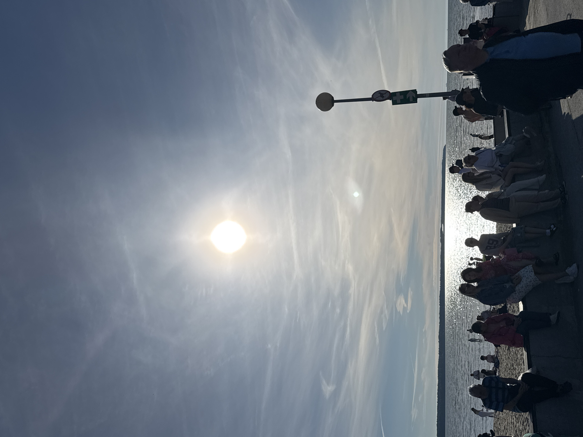 A group of people are seated on a promenade beside a body of water, possibly a bay or a lake, with the sun shining brightly in a clear blue sky with wispy clouds. The sun is positioned in the upper left quadrant of the image, casting a strong glare. The people are facing towards the water, some sitting on a ledge, others on a sandy area. Some appear to be looking out at the water, while others are interacting with each other. The scene suggests a relaxed, leisurely atmosphere, possibly a popular spot for people to gather and enjoy the day.
In the background, there is a street lamp with a sign that appears to have a circular red sign with a line through it, indicating a prohibition, and a green sign with a white arrow, suggesting direction. The water's surface is glittering with sunlight. The overall impression is of a pleasant day out in a public space. The time of day appears to be late afternoon or early evening, given the angle of the sun. The weather is clear and sunny. The location context is Tallinn, Estonia. No discernible text is visible in the image besides the signage on the street lamp.
