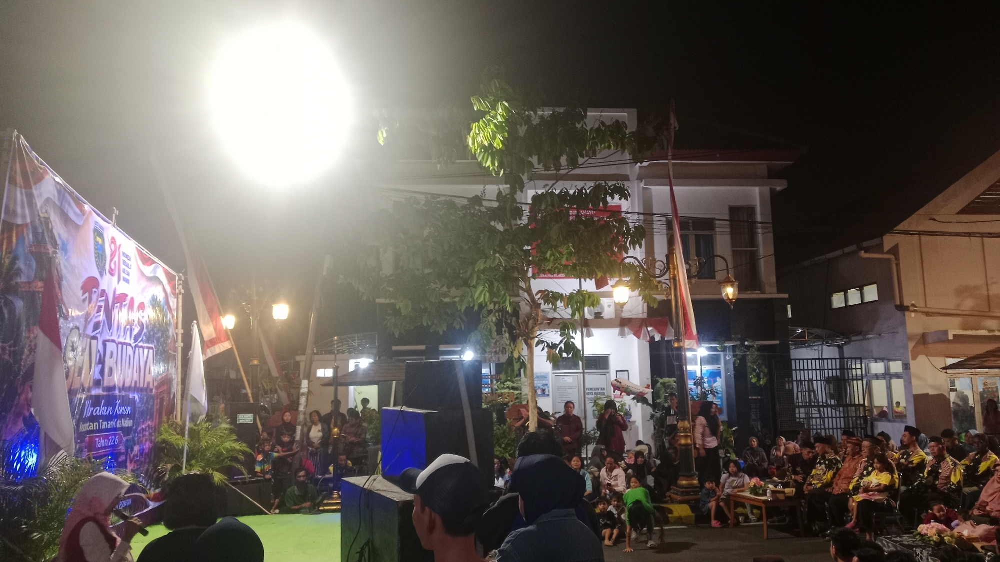 The image captures a nighttime outdoor event in Madiun, Indonesia, likely a cultural performance or festival. A large banner, partially illuminated by a bright spotlight, is prominent on the left, displaying text in Indonesian, including "PENTAS SENI BUDAYA" (Cultural Art Performance) and "KIRAHA KUNCEN" (Kuncen's Jewel). Several Indonesian flags are visible, including a red and white flag. A crowd of people are gathered, some seated on the ground or on low stages, while others stand and observe. A stage area appears to be set up with speakers and lighting equipment. The background shows a building with windows and an illuminated sign. The atmosphere suggests a community gathering celebrating local arts and culture.