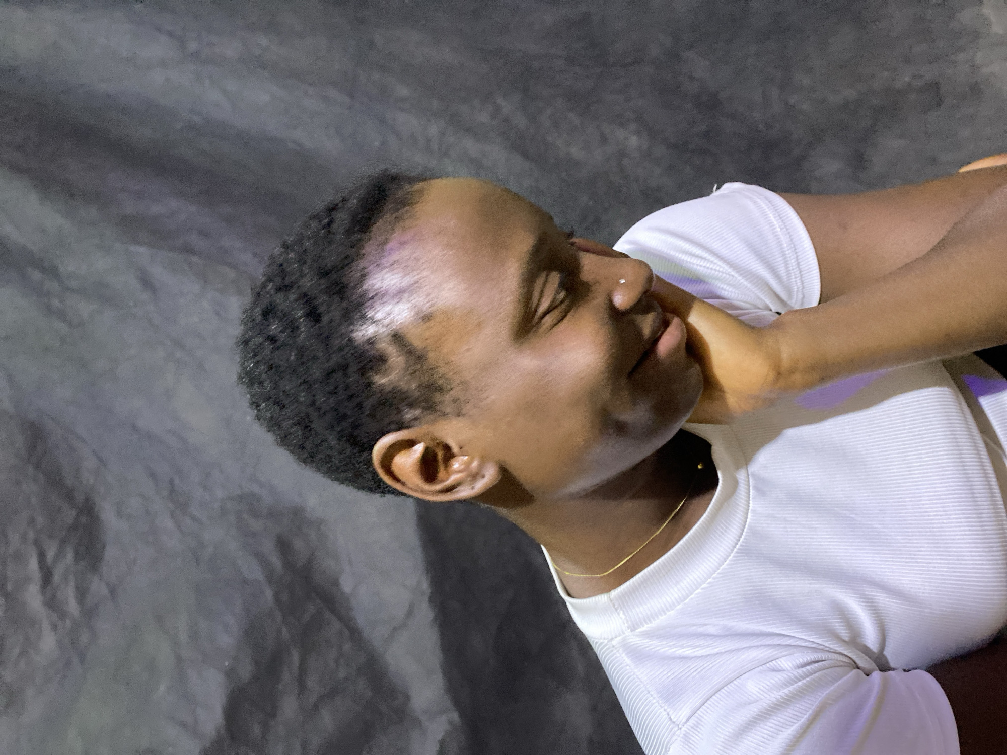 An individual, likely female, with a dark complexion and short, dark, curly hair, is captured in a close-up, medium shot. The person is reclining or lying down, with their head tilted upwards and eyes gently closed, suggesting a moment of peace, rest, or contemplation. Their right hand is softly resting against their right cheek and jawline.
They are wearing a fitted, white, short-sleeved top with a ribbed texture. A thin, delicate gold chain necklace is visible around their neck. A small, shiny stud piercing can be seen on their right nostril. On their forehead, near the hairline, there is a distinct patch of light purple or iridescent sheen, possibly a reflection from lighting, a cosmetic application, or a skin variation. The intricate details of their right ear are also clearly visible.
The background is a dark, mottled fabric or surface, possibly a draped cloth or blanket, which provides a stark contrast to the white top and the subject's skin tone. The lighting appears to be focused on the subject, creating highlights on their face and clothing against the dim background. No specific location cues are visible within the image itself, other than the provided context of Kuru Dungus, Nigeria. There is no visible text in the image. The overall mood conveyed is one of tranquility and calm.
