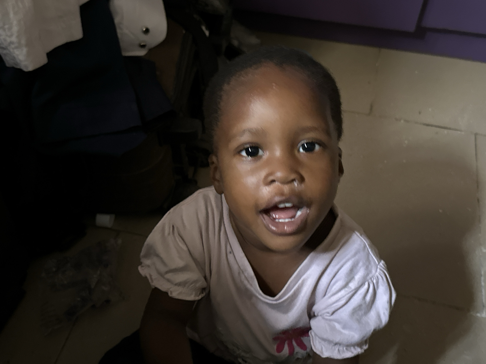 A young Black child, appearing to be a toddler, is seated on a tiled floor, looking directly at the camera with an open mouth and wide eyes. The child is wearing a light pink t-shirt with puffed sleeves and a floral design on the front. The background is dimly lit and appears to be an indoor setting, possibly a room in a house. To the left of the child, dark fabric is visible, and in the background, a purple surface and a wall are discernible. The floor tiles are a neutral color. The scene suggests a casual, domestic environment within Narayi, Nigeria.